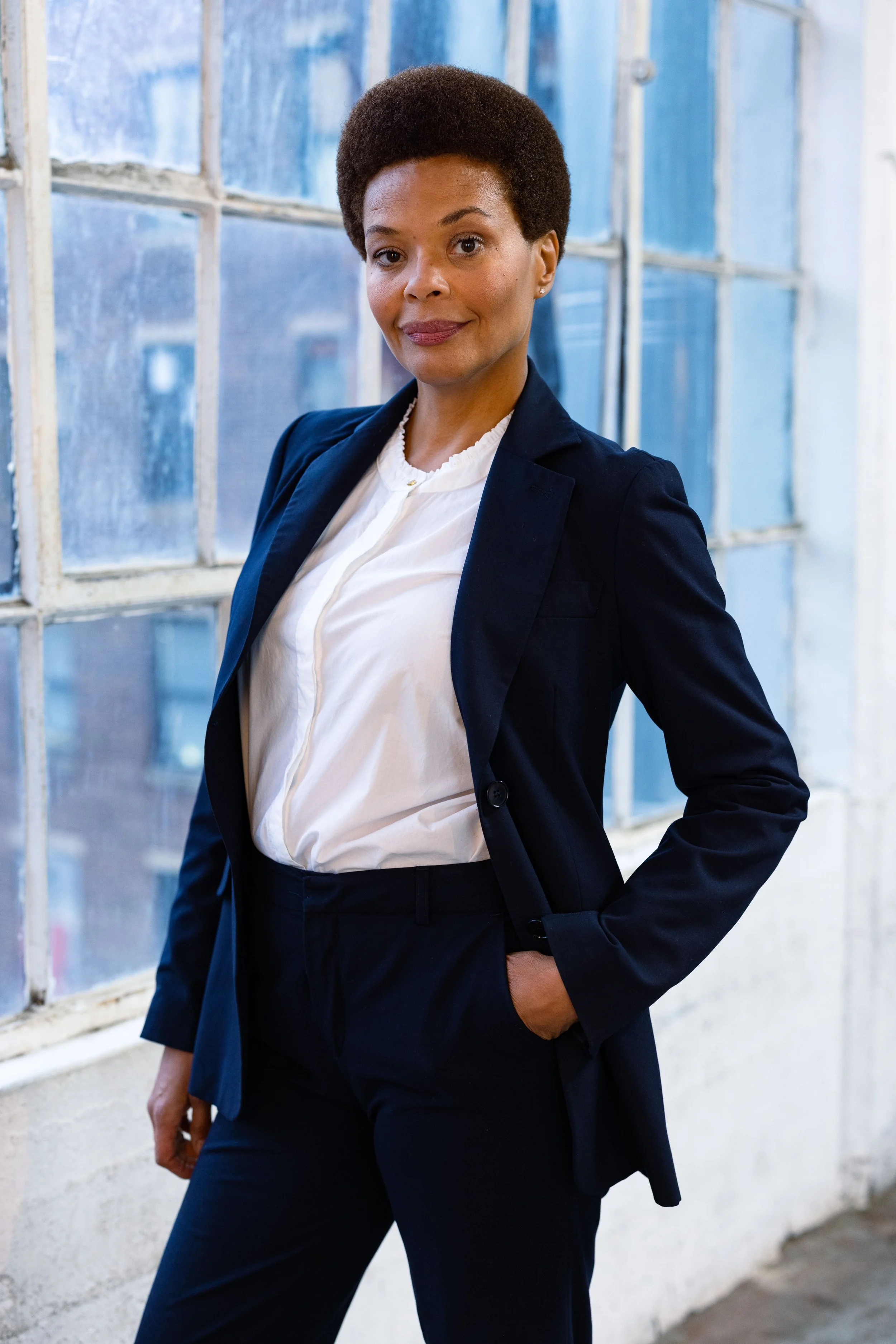 A confident woman in a navy blazer and white shirt standing by a large industrial window.