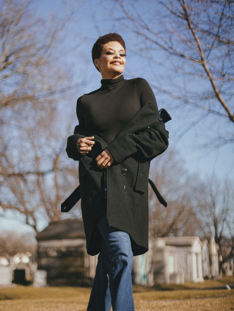 A woman, Erickka Jones, with short curly hair smiling outside on a clear day, wearing a black turtleneck, a black coat, and blue jeans, with bare trees and a park-like area in the background.