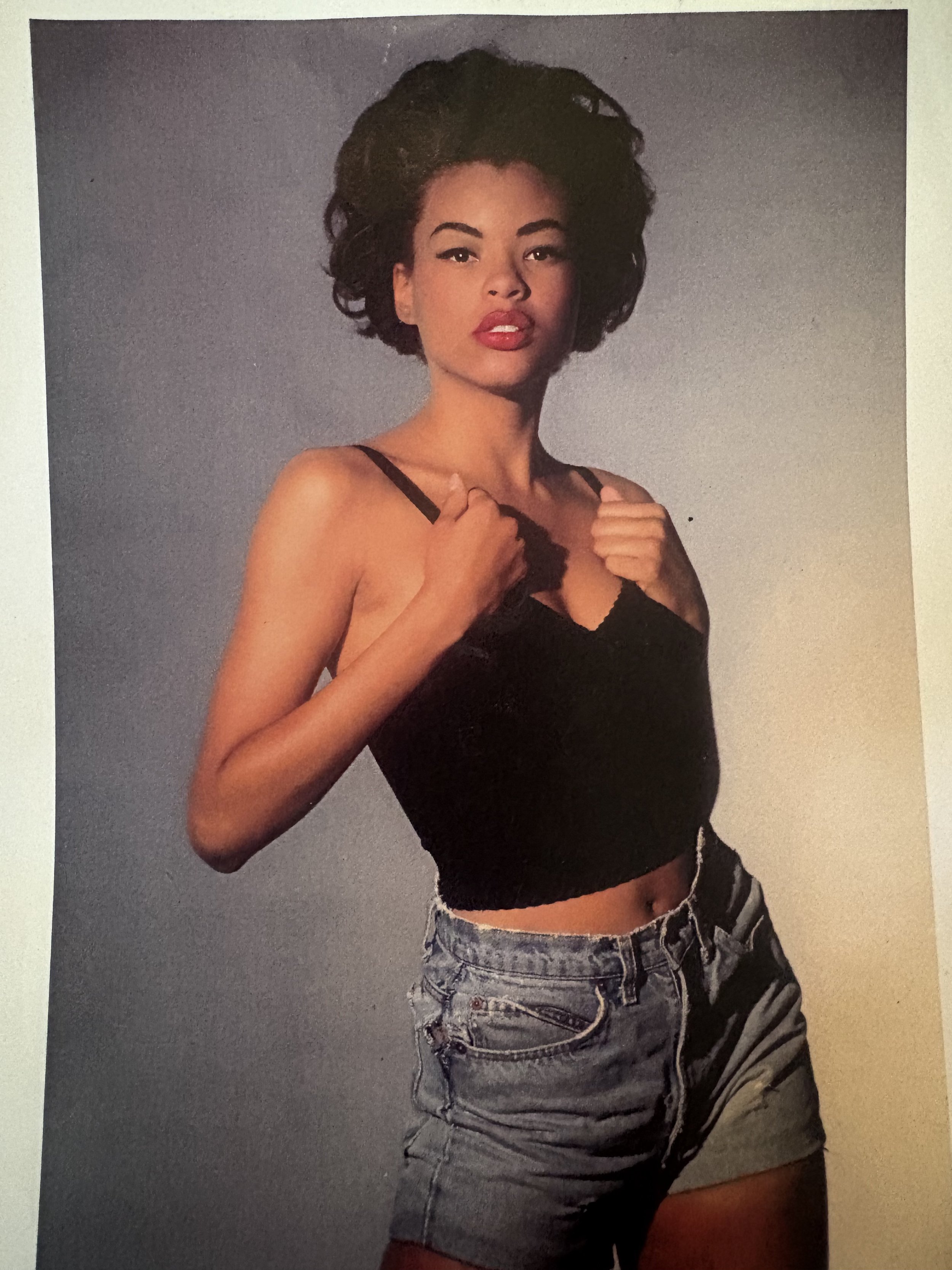 A young Erickka Jones with curly dark hair, red lipstick, and a confident expression wearing a black spaghetti strap top and high-waisted denim shorts, standing against a plain background.