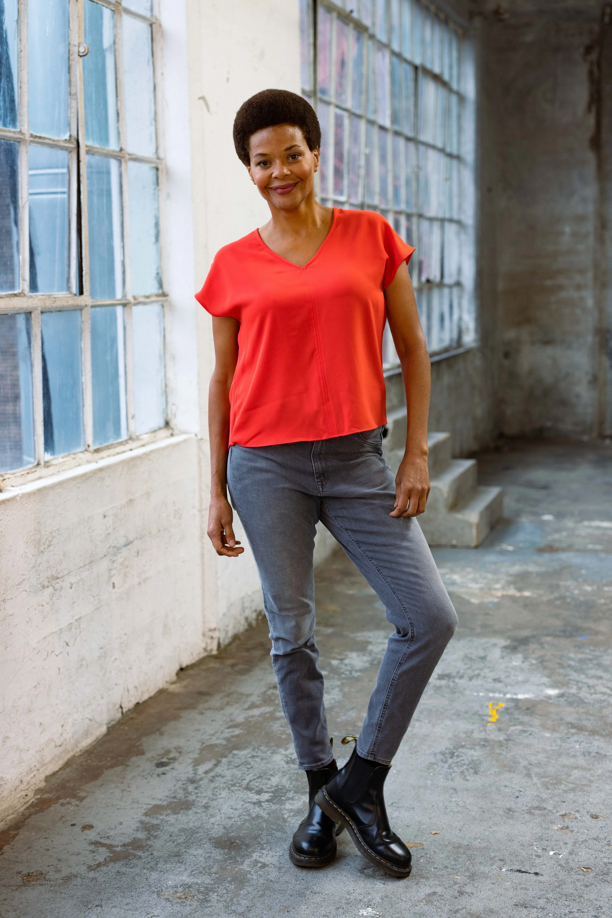 A woman with short curly hair and dark skin wearing a red blouse, gray jeans, and black ankle boots stands in front of a large window in an industrial-style space with concrete floors and walls.