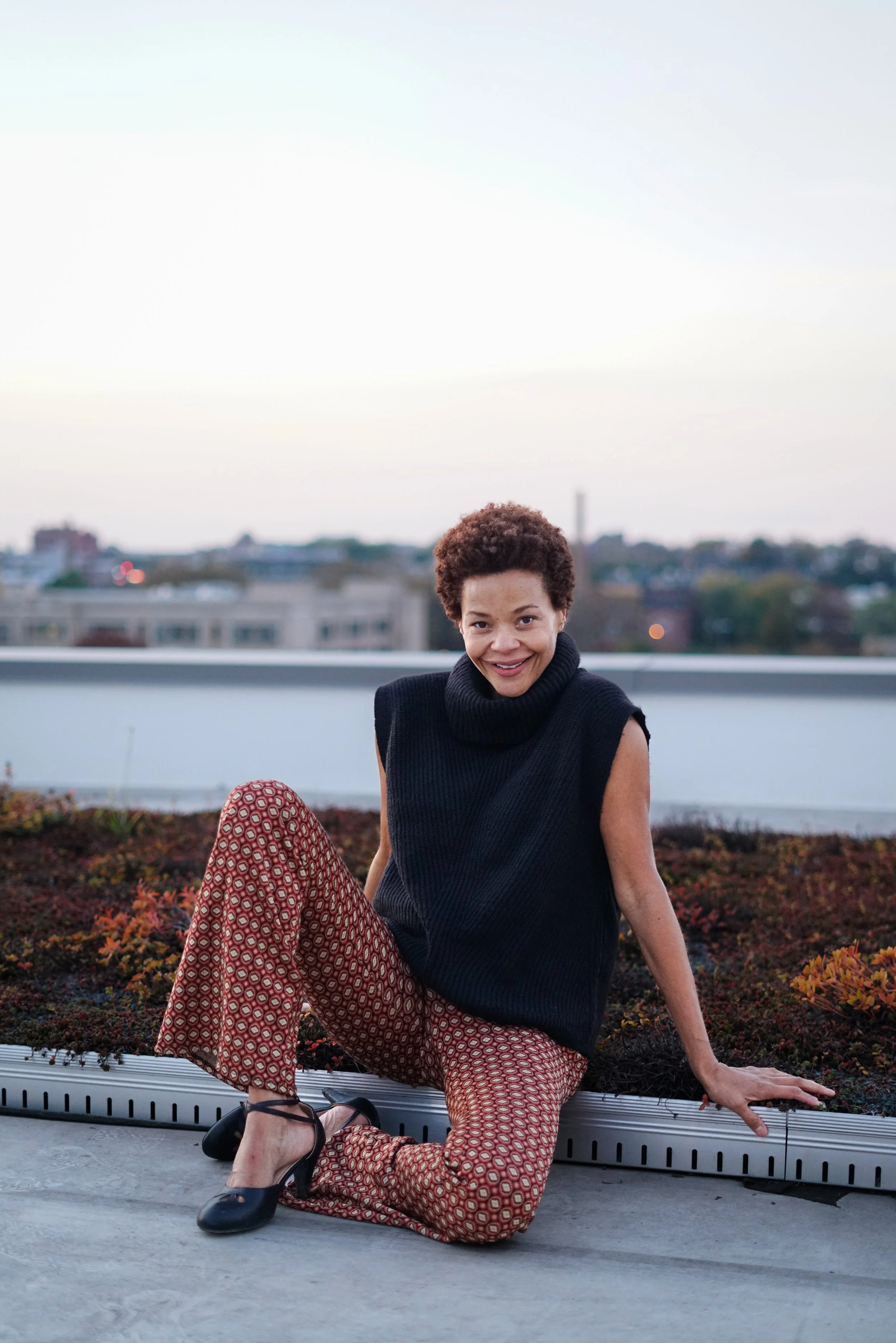 Woman with curly hair sitting outdoors on a rooftop, smiling, wearing a black sleeveless top, patterned red and beige pants, black heels, against a blurred cityscape background at dusk.