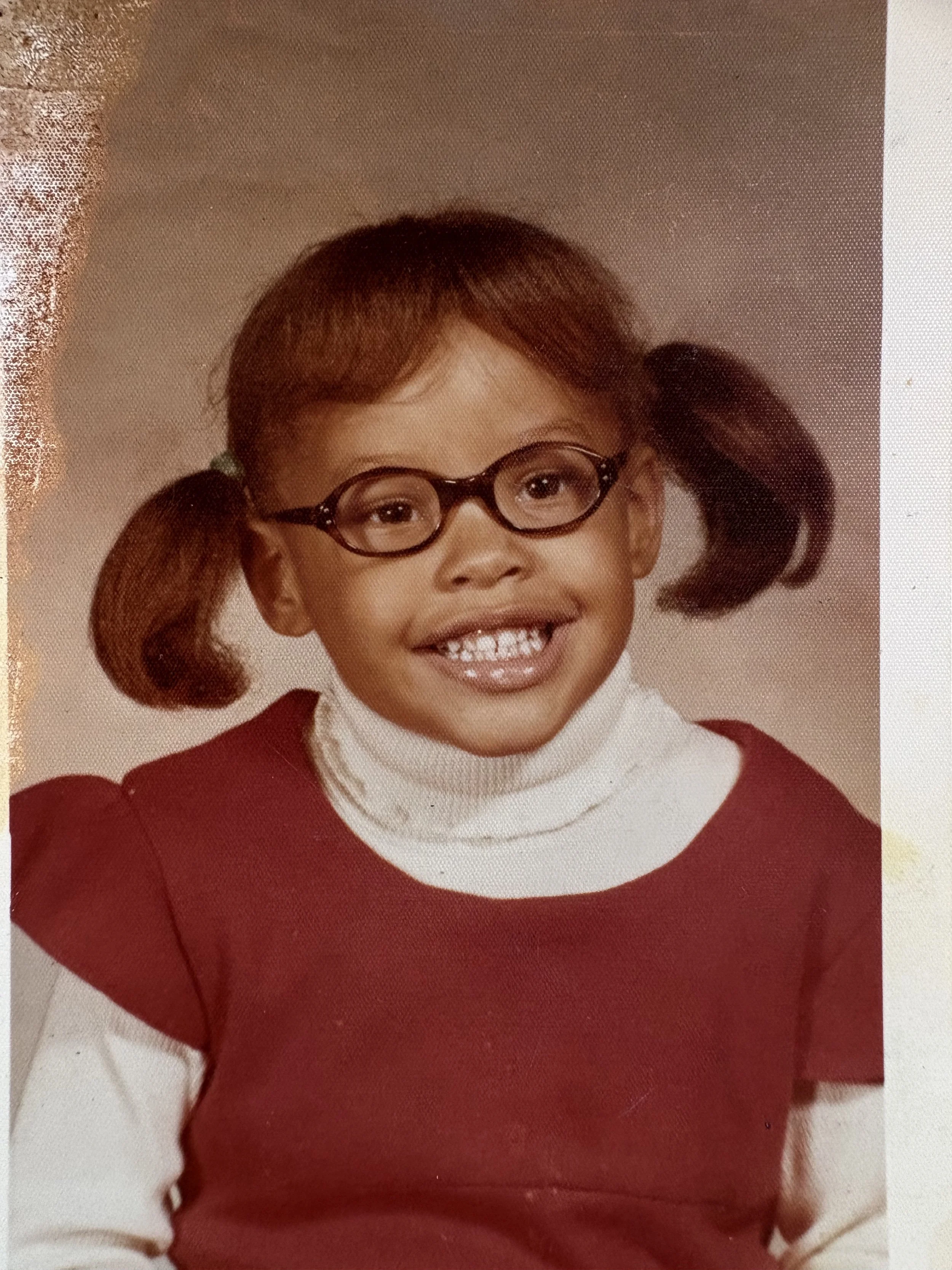 A young Erickka Jones with glasses, pigtails, wearing a red sweater over a white turtleneck, smiling for a school photo.