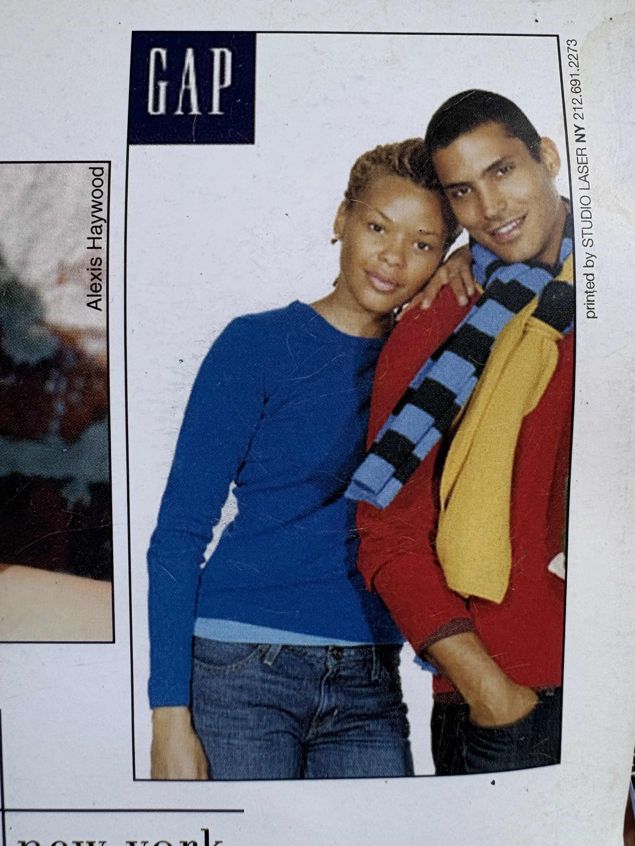 Erickka Jones with blonde locs wearing a blue long-sleeve shirt and a man with short dark hair wearing a multicolored jacket, blue and black checkered shirt, and yellow shirt, posing together with their arms around each other for a GAP ad. 