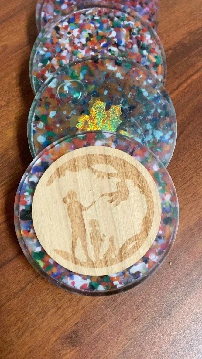 Four round containers filled with multicolored glitter and confetti, with the bottom container featuring a stencil of a person shaking hands with a frog, and a leaf above them.