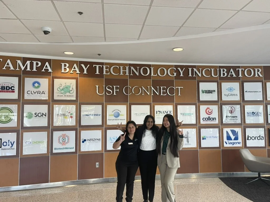 Three women standing in front of a wall with business logos at Tampa Bay Technology Incubator, USF Connect, smiling and posing for the photo.