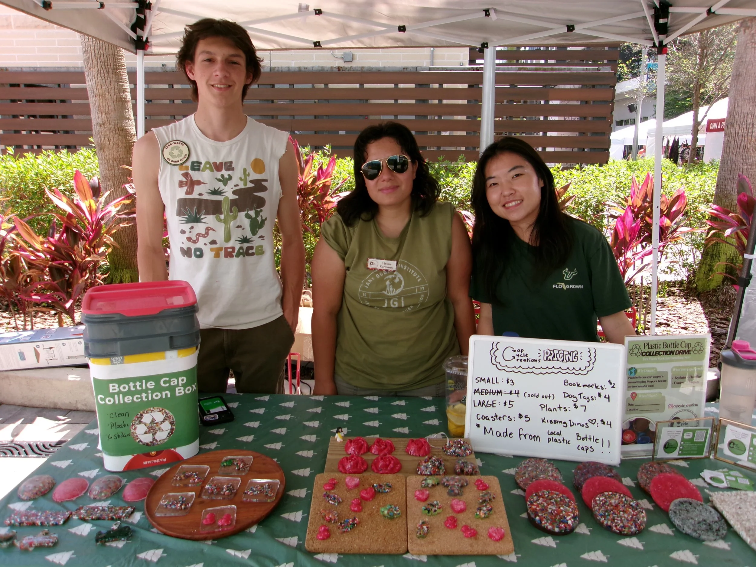 Three people standing behind a table with various decorated cookies at an outdoor event. The table features a sign about bottle cap collection and pricing.