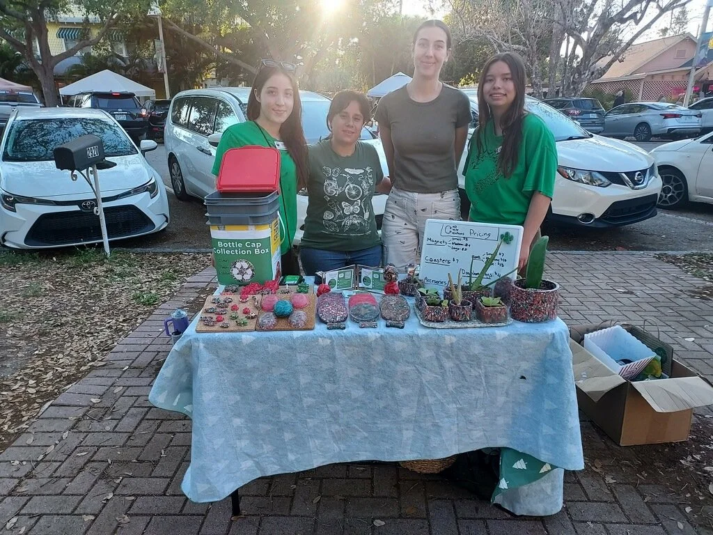 Four people standing behind a table with handmade crafts and a sign, outdoors in a parking lot lined with cars, trees, and tents.