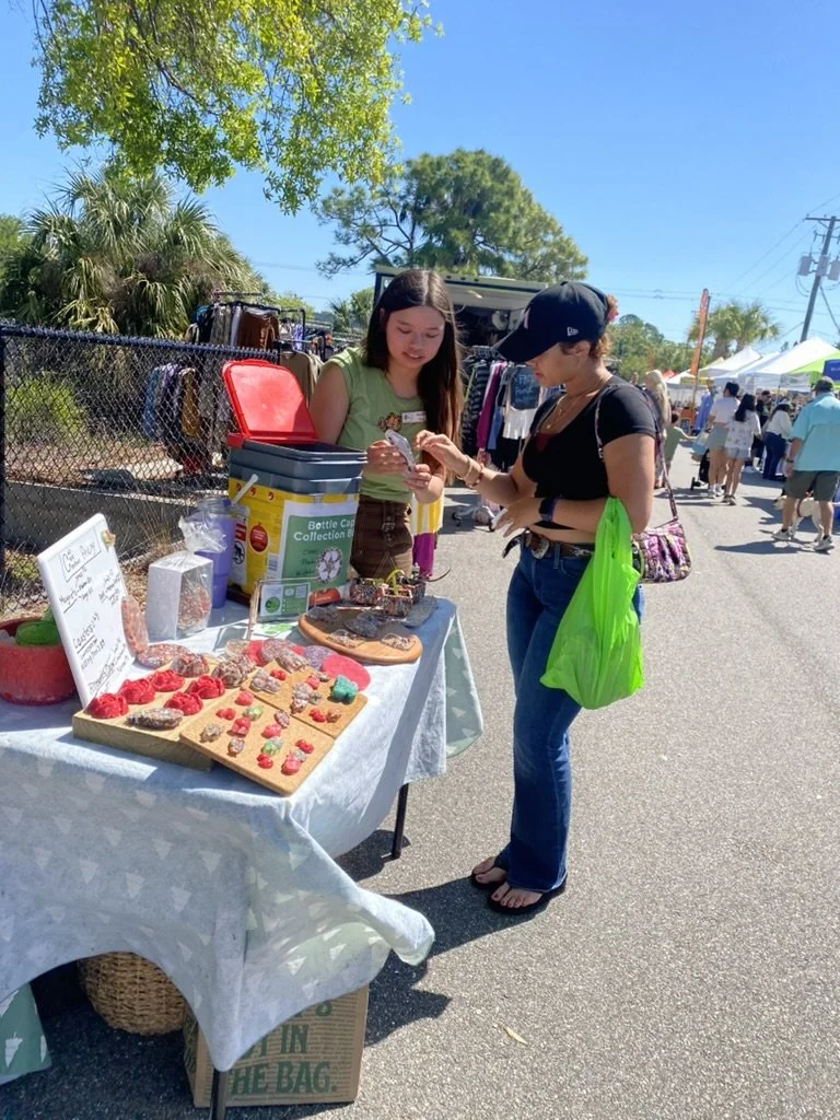 Two women are standing at a vendor stall at an outdoor market. The stall has small, colorful handmade crafts, possibly miniature figurines. One woman is handling money while the vendor woman is accepting it. The setting is a sunny day with groups of people walking in the background.