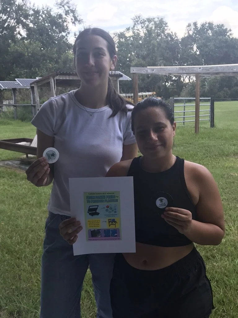 Two women standing outdoors in a grassy area, holding small round objects with a logo, and a poster with environmental messages. One woman is wearing a white shirt and the other a black crop top.