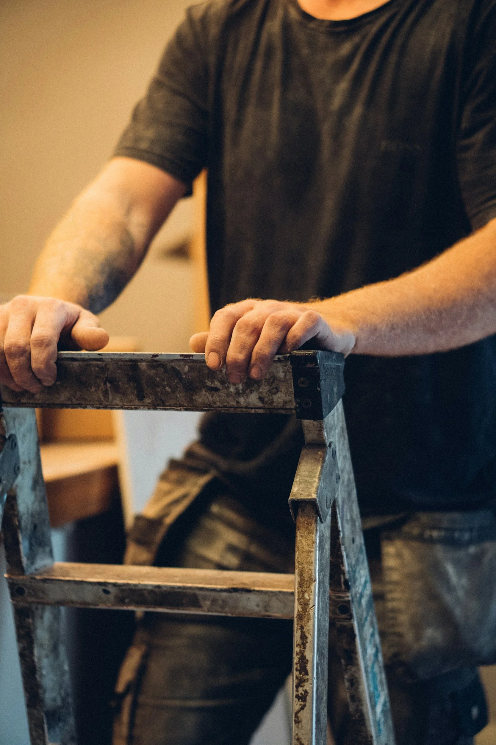 Close-up of a person wearing a black t-shirt and camouflage pants, standing behind a worn, metal stepladder with their hands resting on the top step.