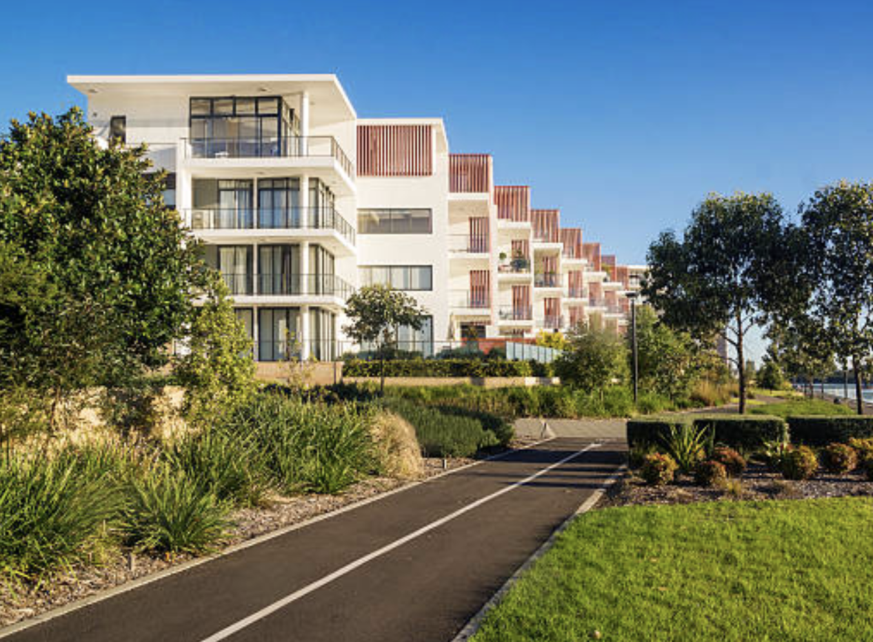 Modern apartment building with balconies, surrounded by greenery and a paved pathway.