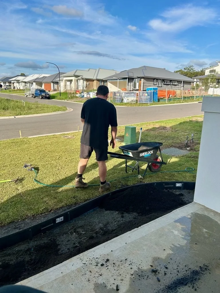 A man working outside on a lawn, with a wheelbarrow labeled 'Sherlock', a green toolbox, and a garden hose, in front of a row of modern houses on a sunny day.