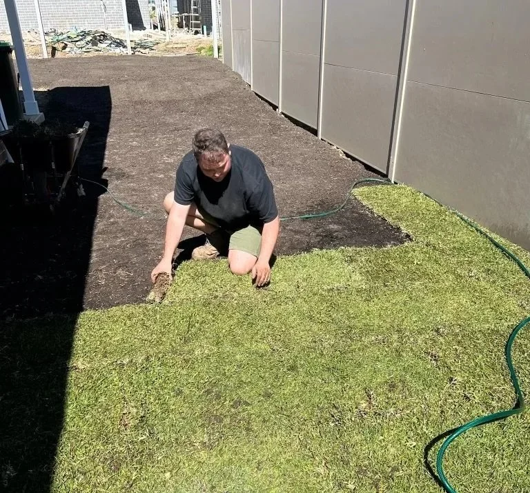 Person laying on the ground in a backyard, planting grass seed or sod between a garden bed and a house wall.