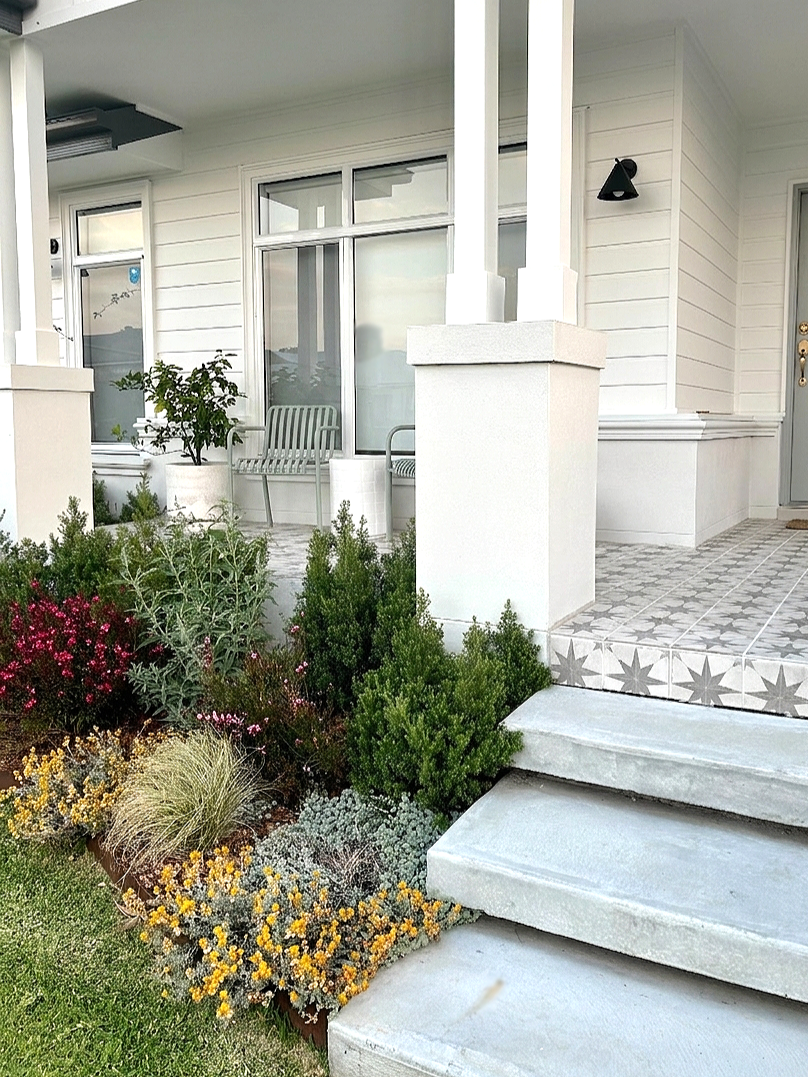Front porch with white siding, decorative tiled steps, black outdoor wall light, and plants and shrubs in the garden.