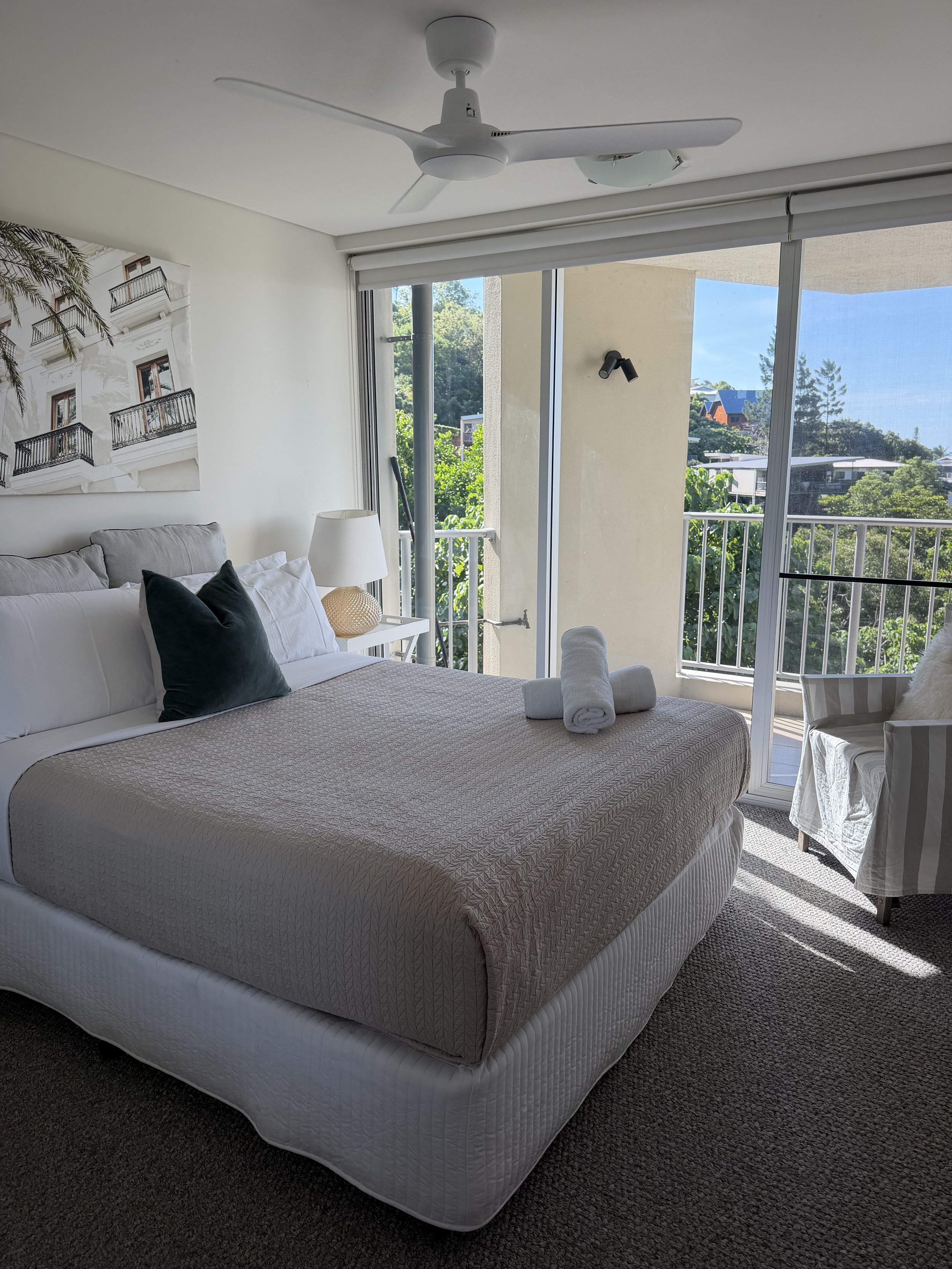 A bedroom with a white bed, black accent pillows, a side table with a lamp, and a balcony with sliding glass doors showing a view of trees and houses.