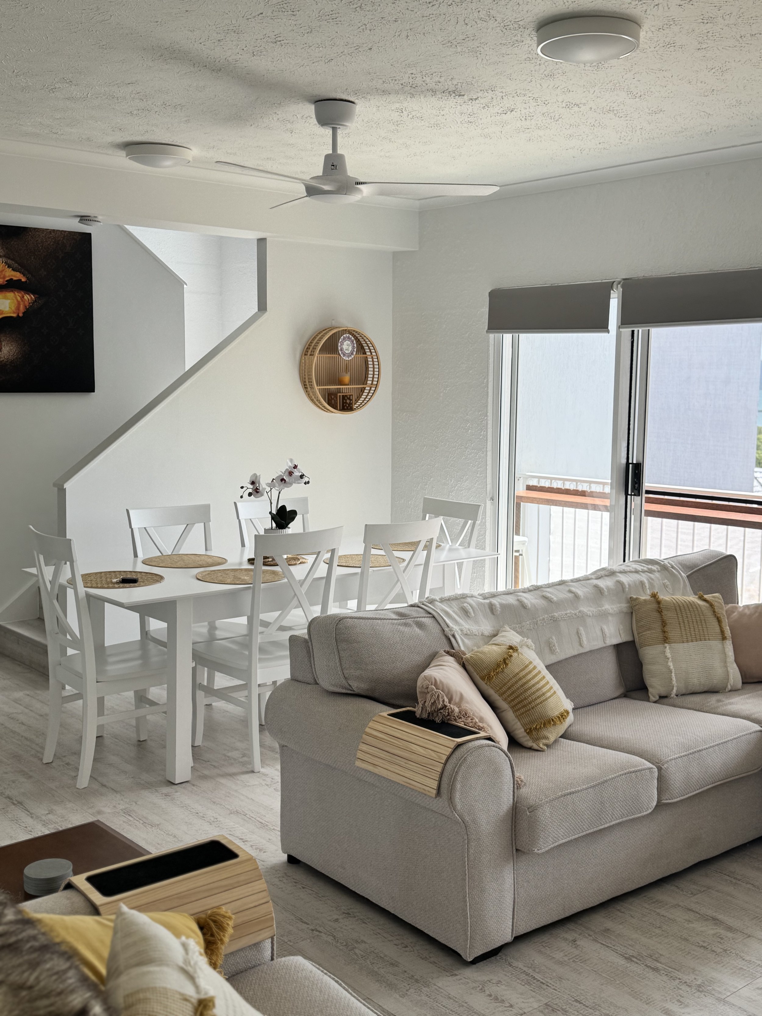 A living room with a white dining table and chairs, a beige sofa with pillows, a sliding glass door with blinds leading to a balcony, and decorative wall shelves, ceiling fan, and lighting fixtures.
