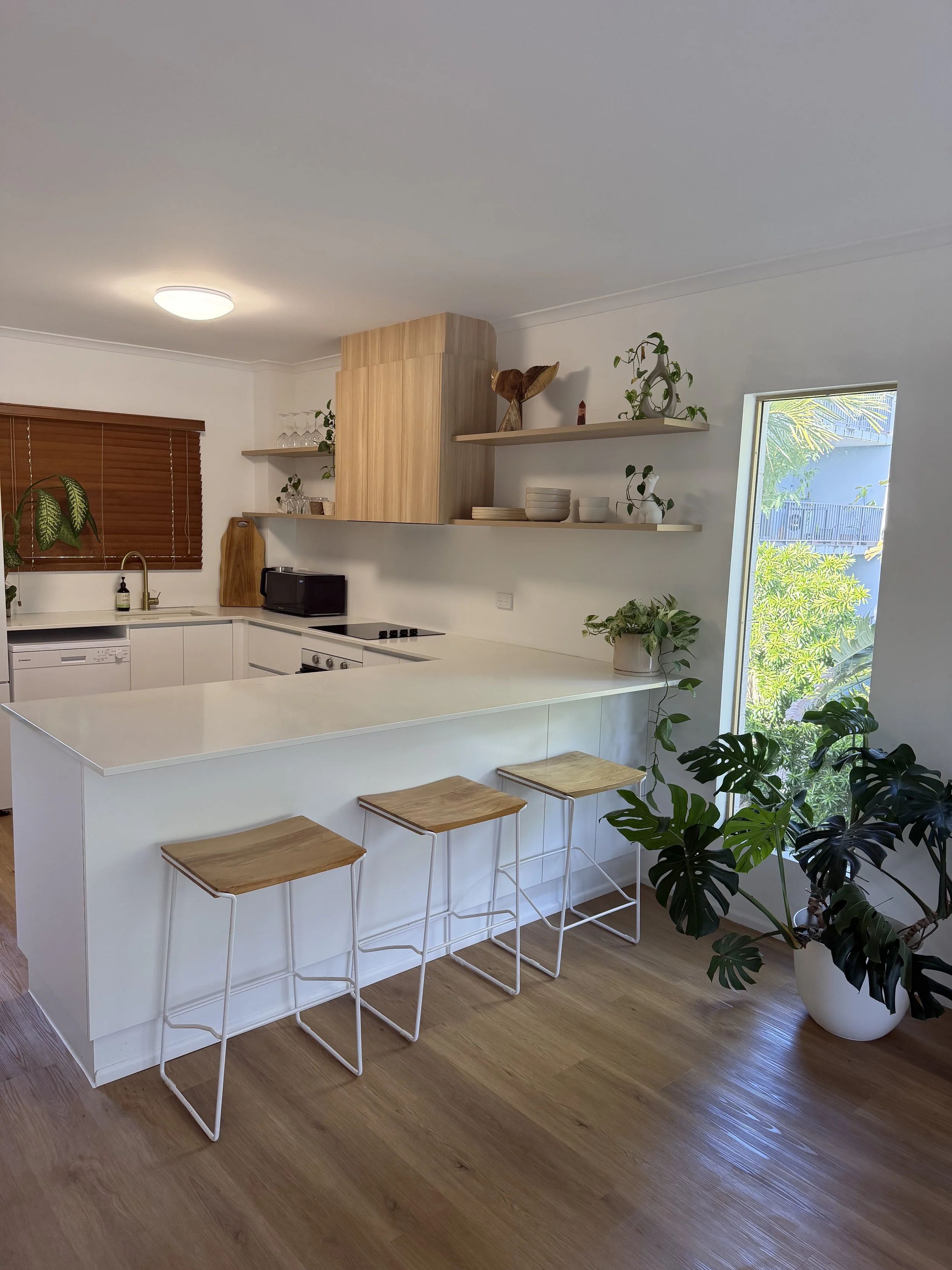Modern kitchen with white countertops, wooden bar stools, wall shelves with plants and decor, large window with greenery outside, hardwood floors, and potted plants.