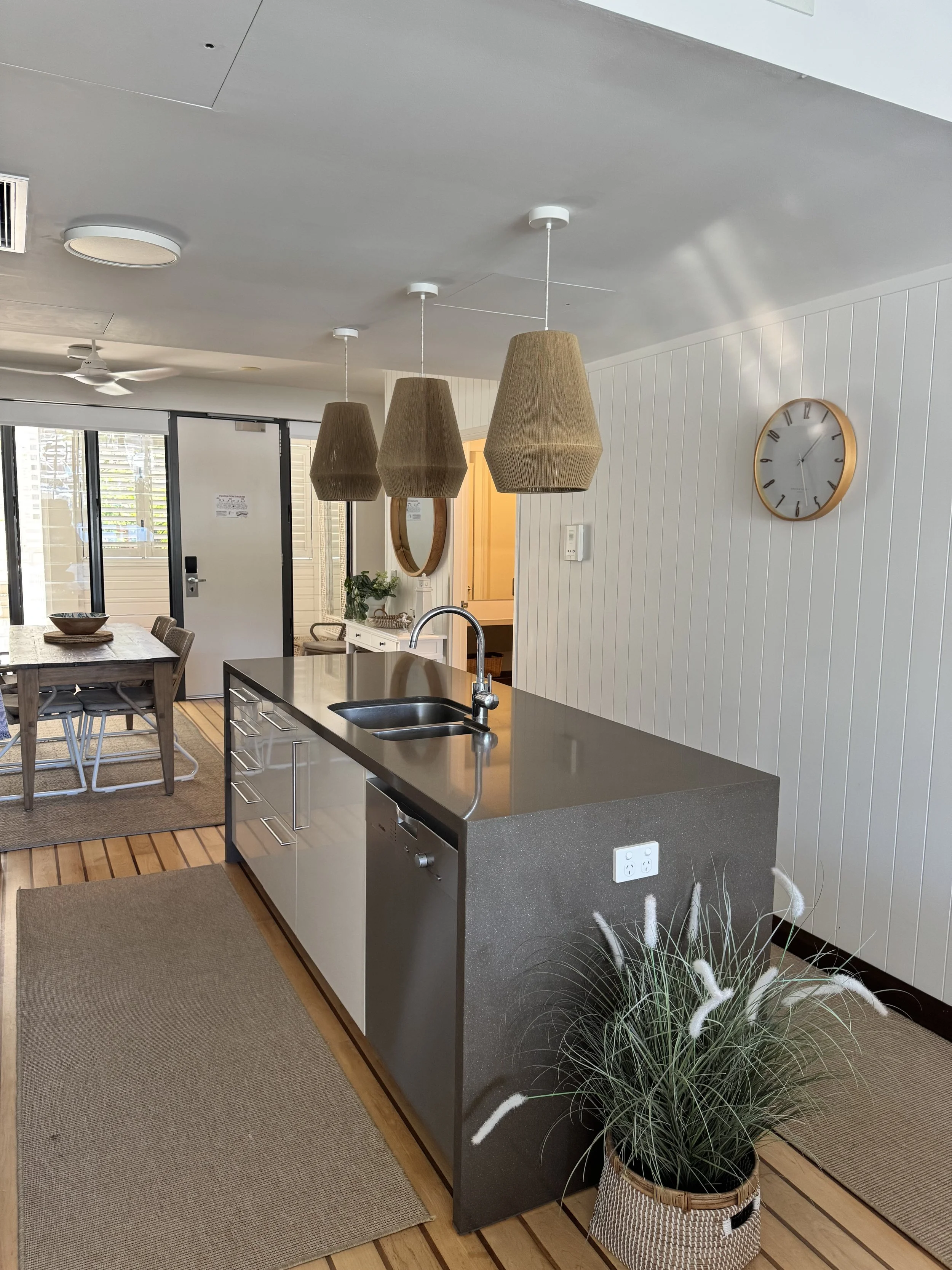 Modern kitchen with pendant lights, a clock on white paneled wall, potted plant on a woven basket, and a sliding glass door leading outside.