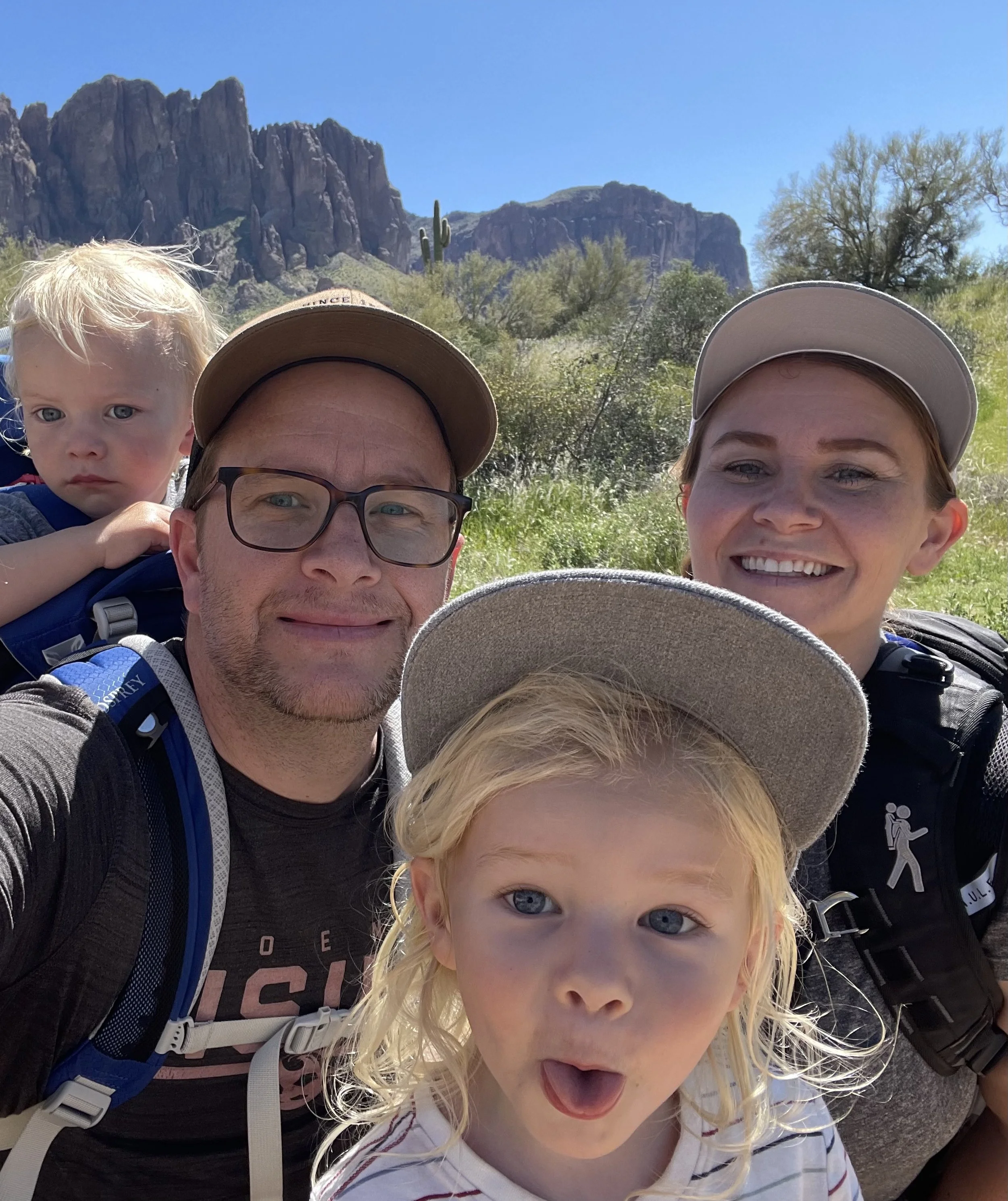 A family of four taking a selfie outdoors with mountains, cacti, and clear blue sky in the background. The group includes a young girl with blonde hair and blue eyes, a woman with brown hair and a hat, a man with glasses and a hat, and a young boy with blonde hair and blue eyes.