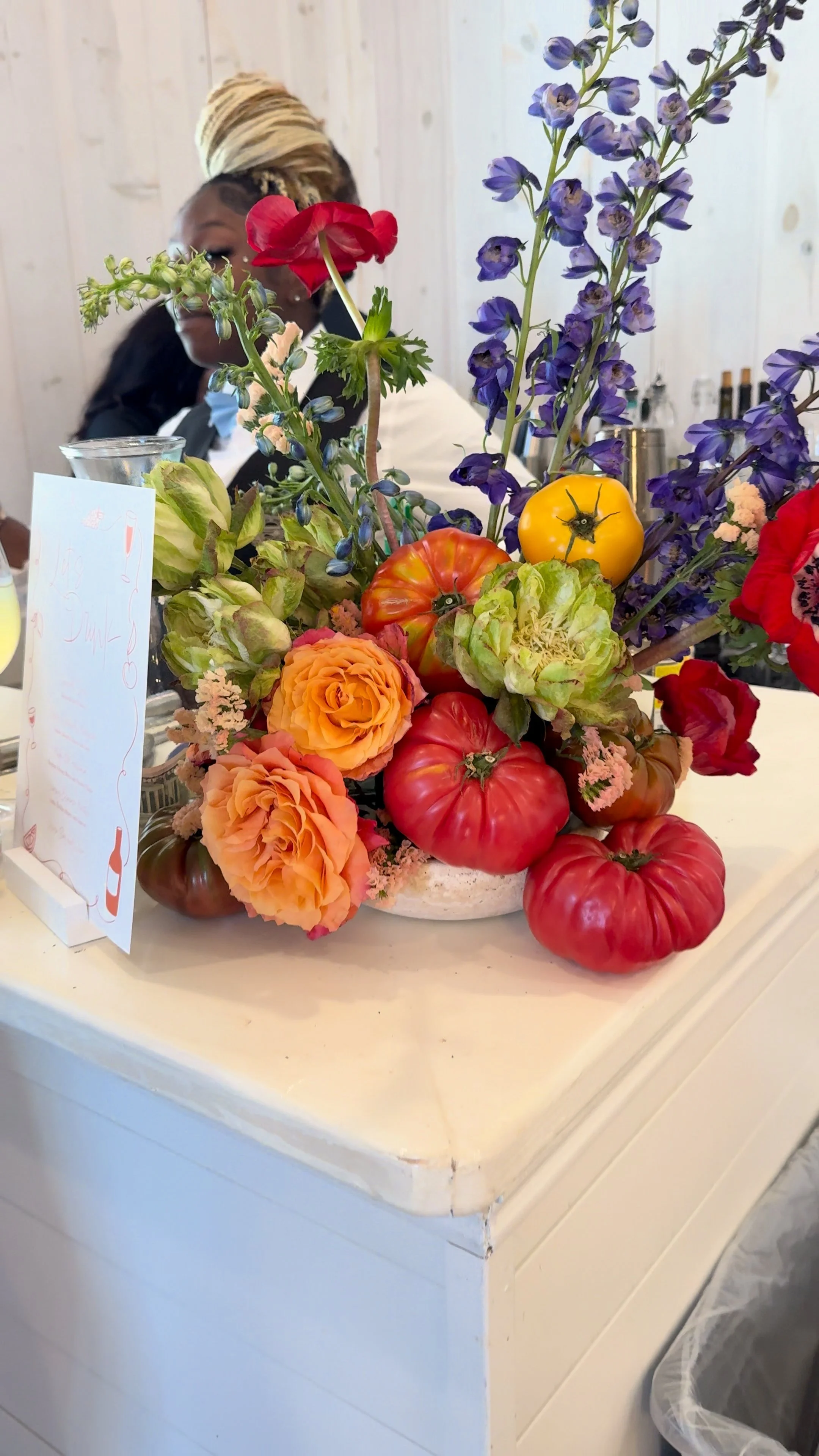 A floral arrangement with red, orange, yellow, purple, and pink flowers and tomatoes on a white surface in a restaurant or cafe setting.
