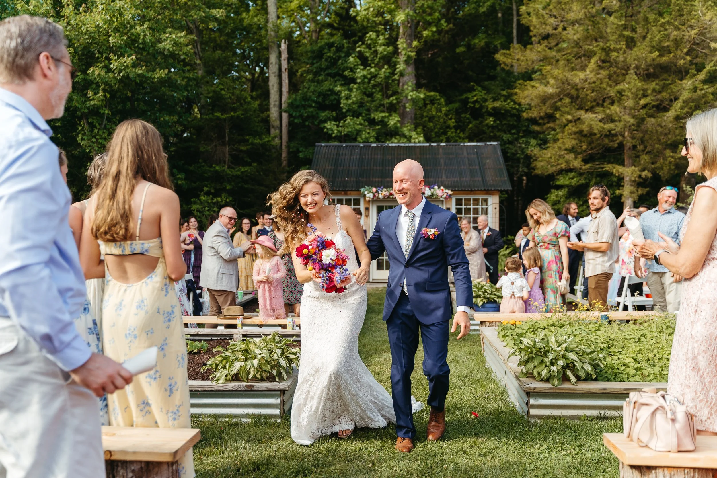 A bride and groom walking together during an outdoor wedding ceremony surrounded by friends and family in a garden with trees and a small building decorated with flowers.