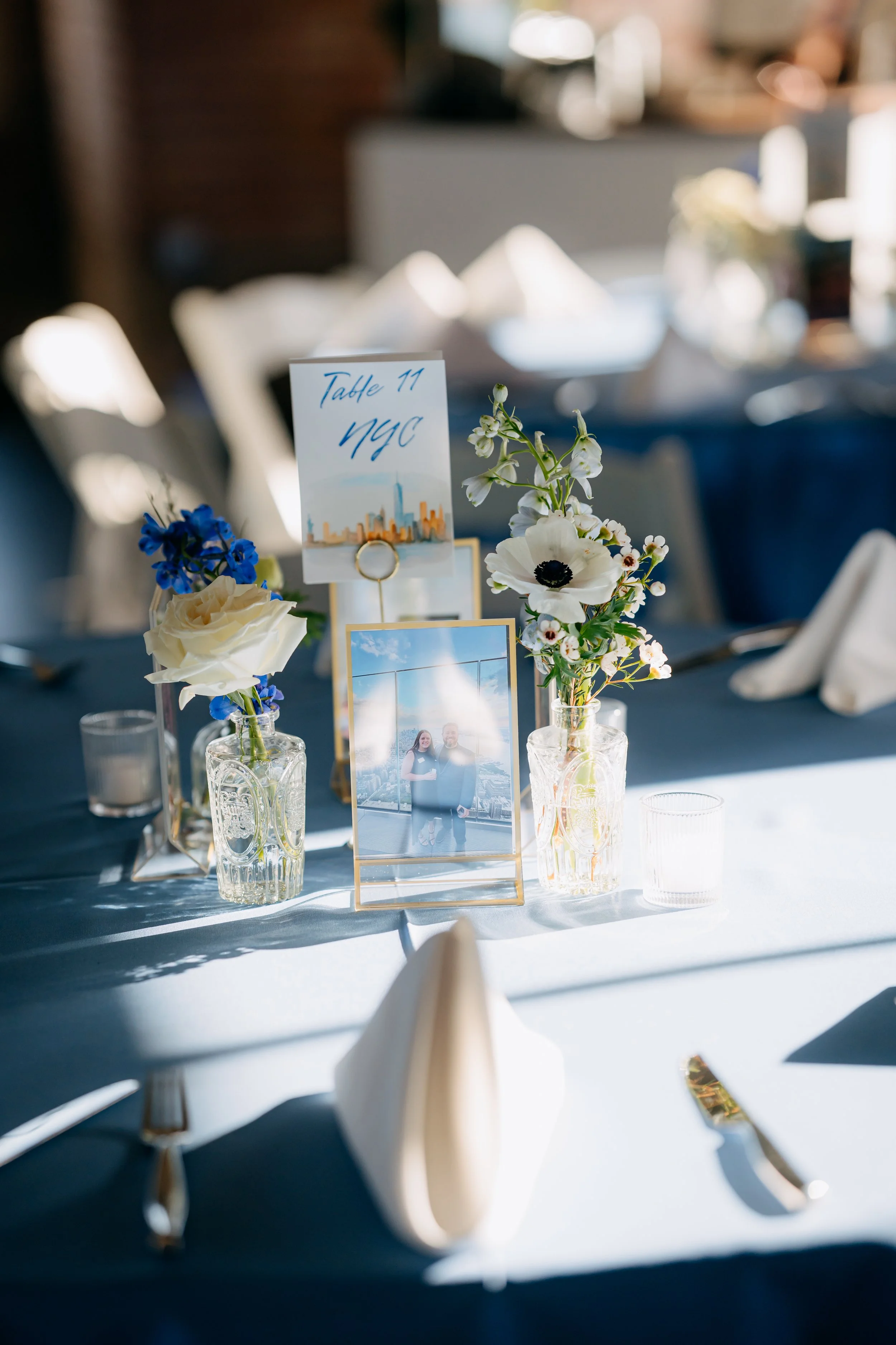 Table decorated with vases of white and blue flowers, framed photos, and a sign indicating "Table 11" at an event or wedding reception.