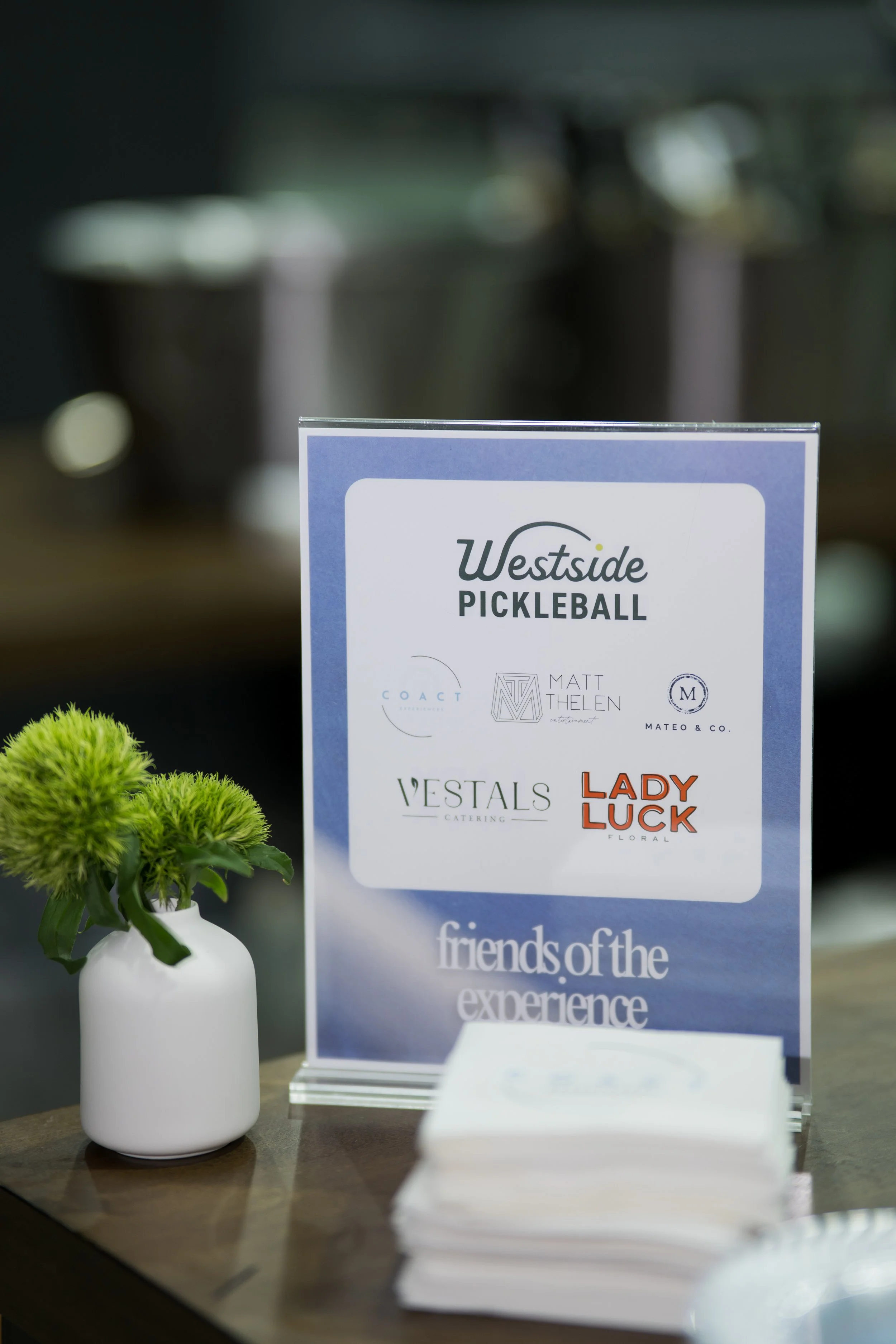 A white vase with a green flower next to a sign that reads 'Westside Pickleball' on a table, with a stack of flyers or brochures in front of it.
