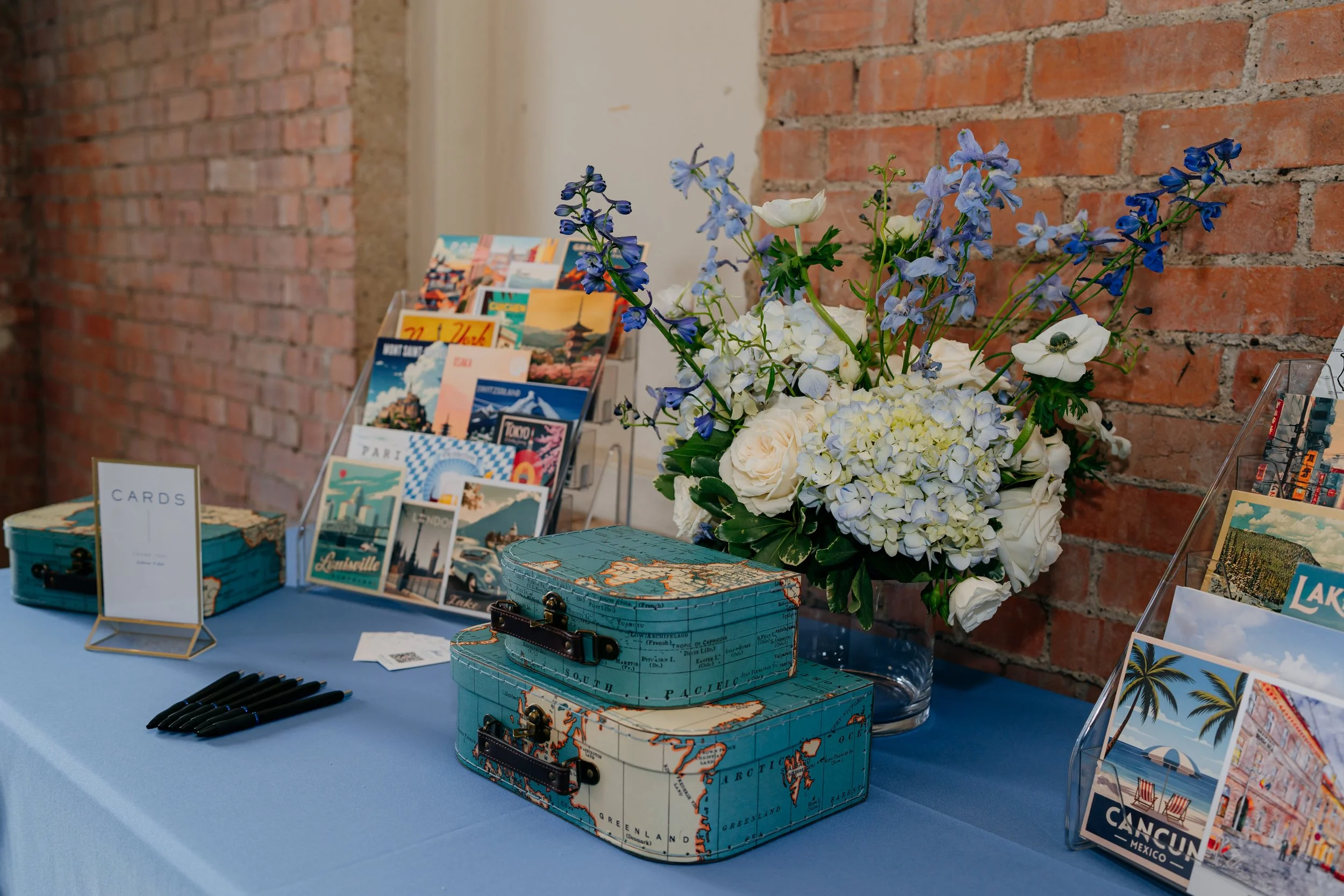 A table displaying decorative boxes shaped like maps, a bouquet of white and blue flowers, and postcards or greeting cards against a red brick wall background.