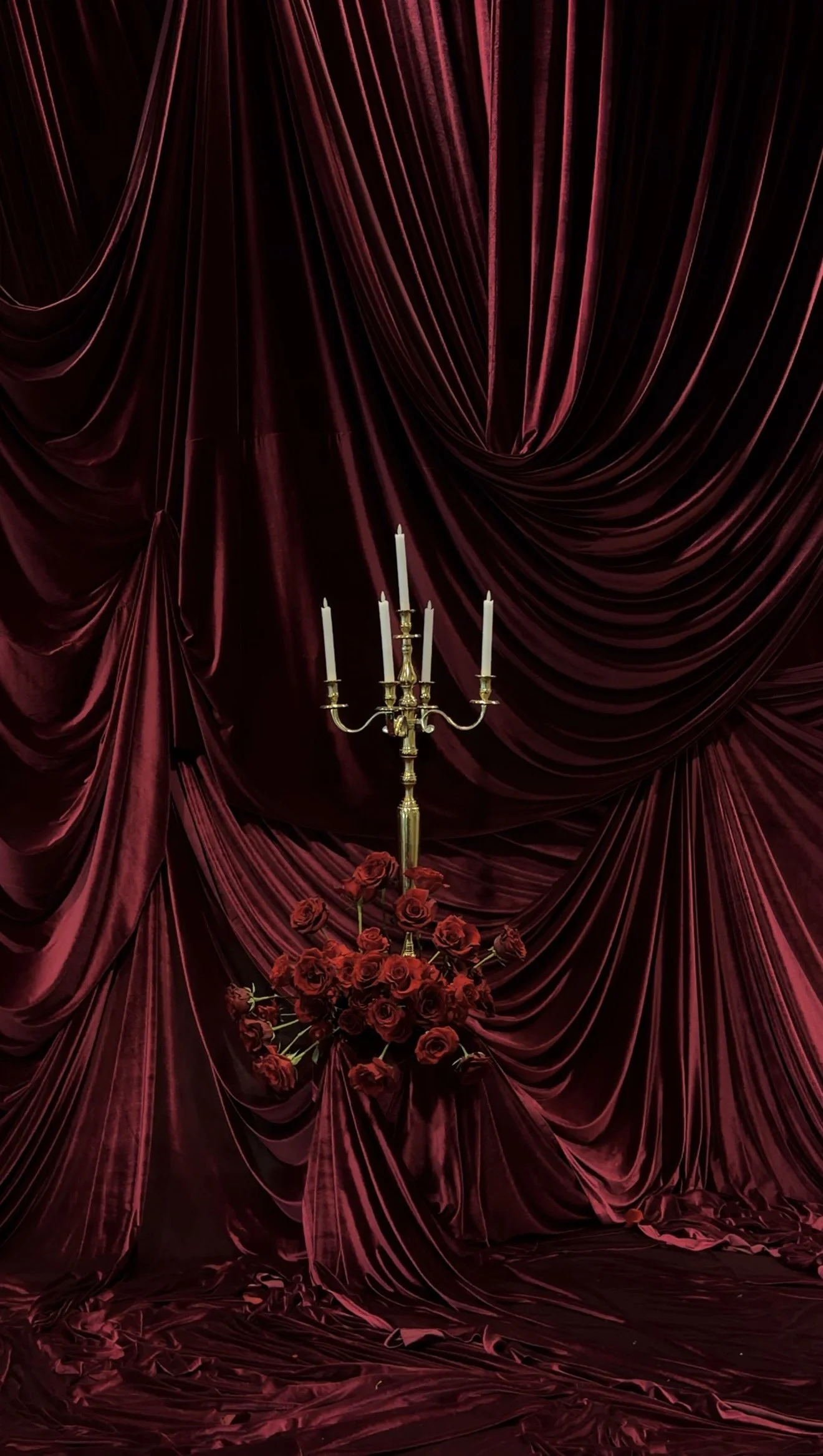 Red velvet curtains with a gold candelabra holding six unlit candles and a large arrangement of red roses in front.