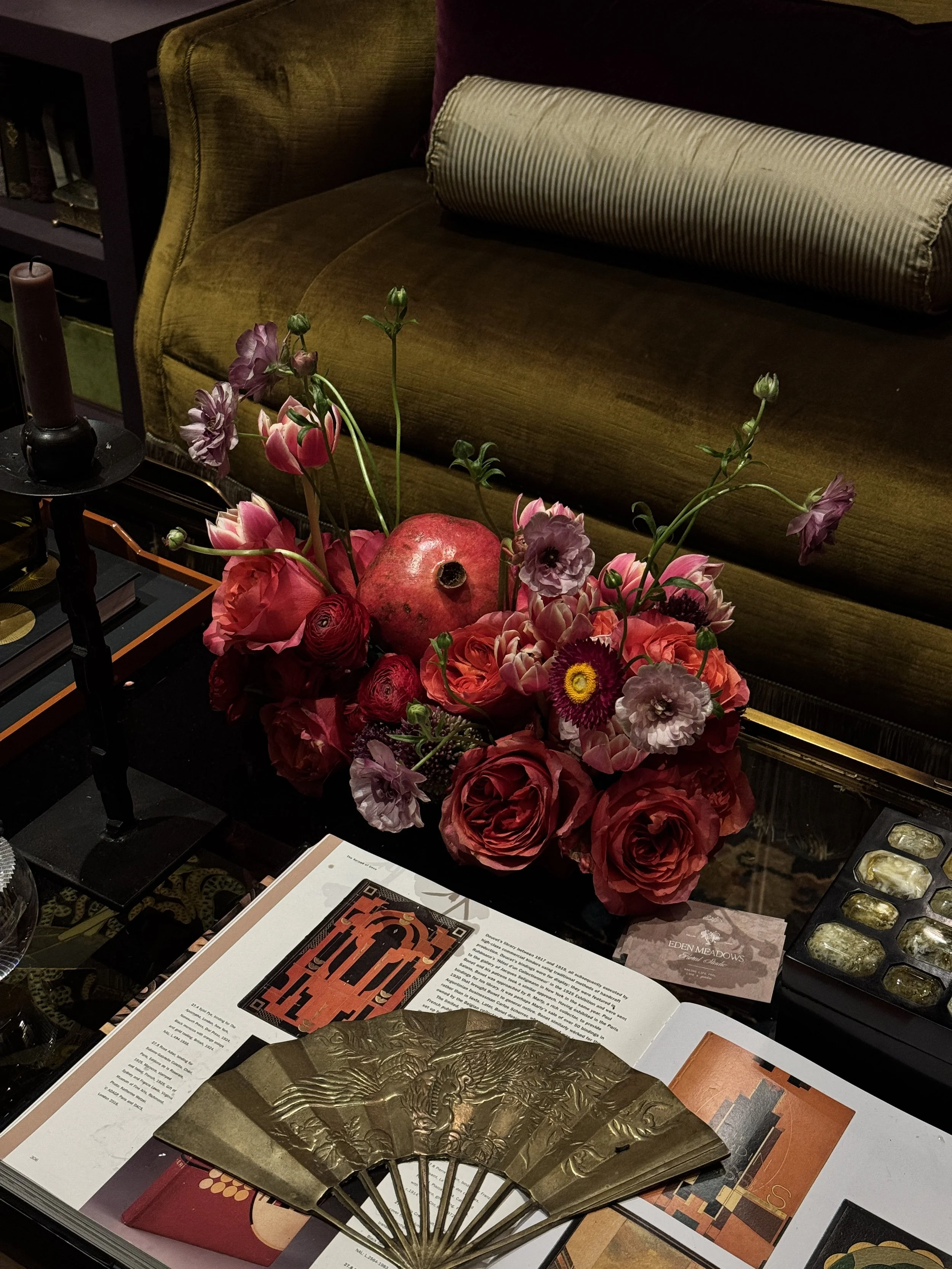 A floral arrangement of pink, red, purple, and yellow flowers placed on a table next to a potted plant, with decorative booklets and a traditional hand fan with gold designs on it.