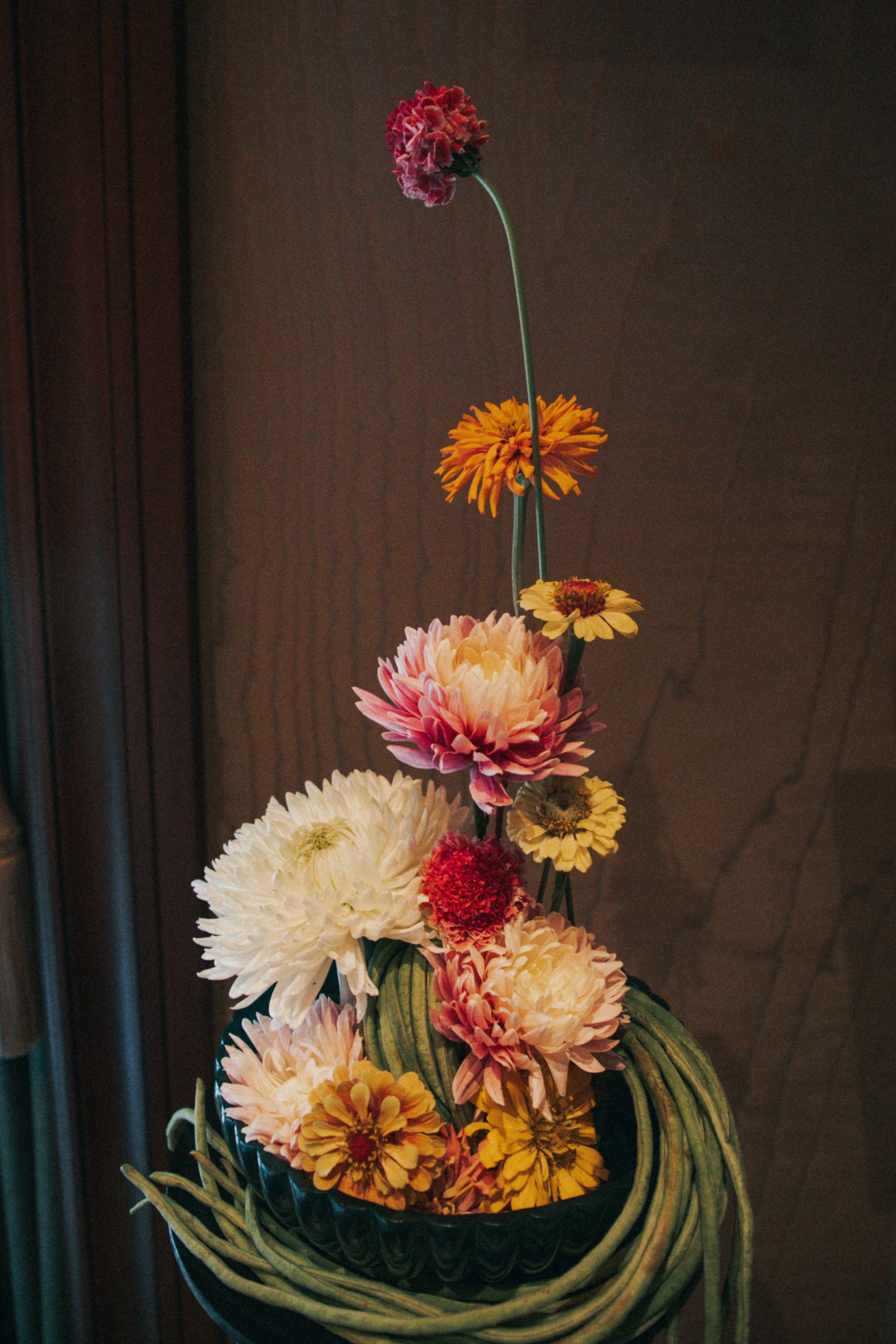 A tall floral arrangement of pink, orange, yellow, and white flowers in a decorative black and yellow ceramic vase on a wooden surface against a dark background.