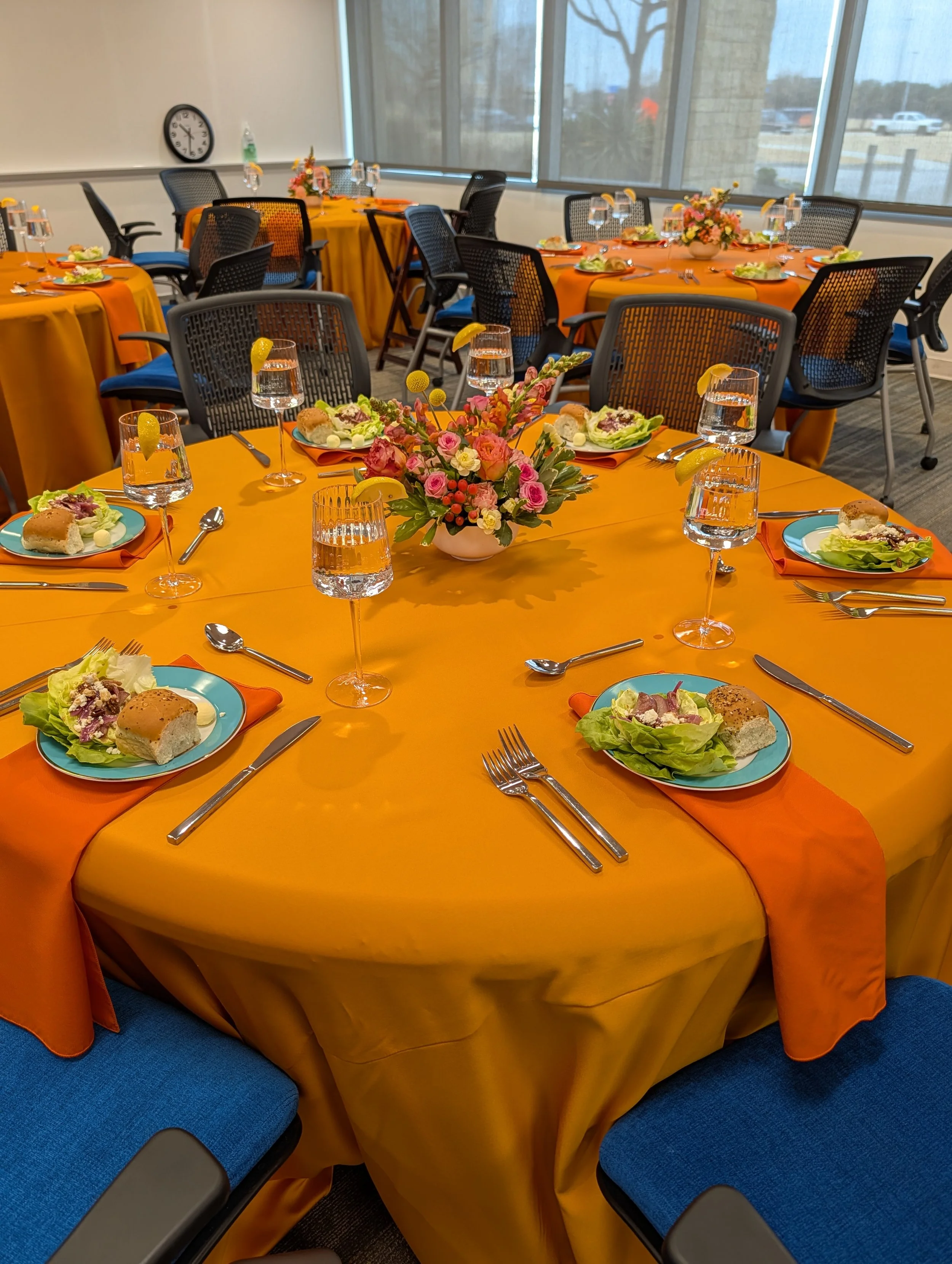 Round tables with orange tablecloths set with salads, bread rolls, lemon slices, and water glasses with lemon wedges. Pink and white floral centerpieces on each table. Blue chairs with black armrests arranged around the tables, in a room with large w