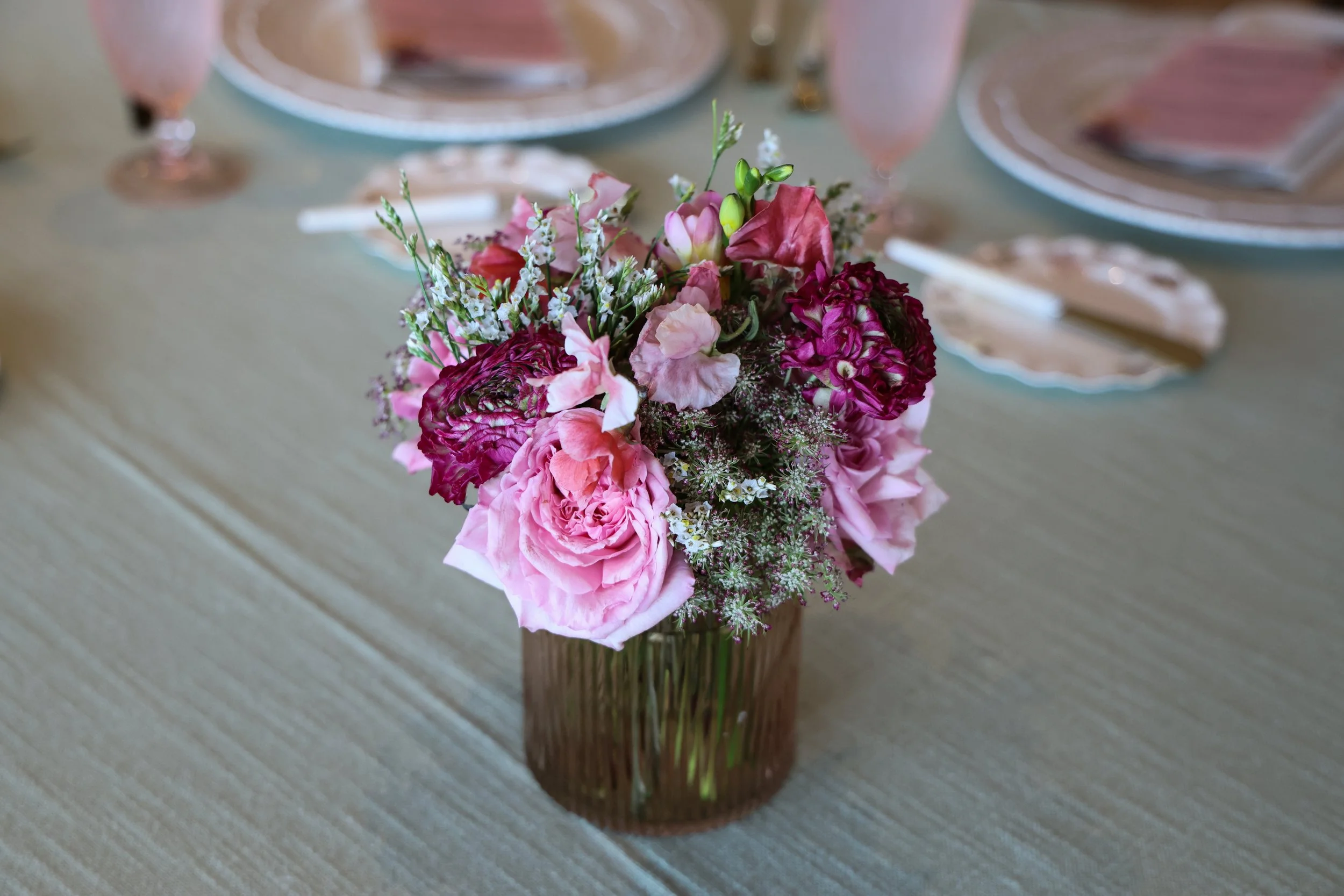 A flower arrangement of pink, purple, and white flowers in a glass vase on a table set for a meal with plates, glasses, and utensils.