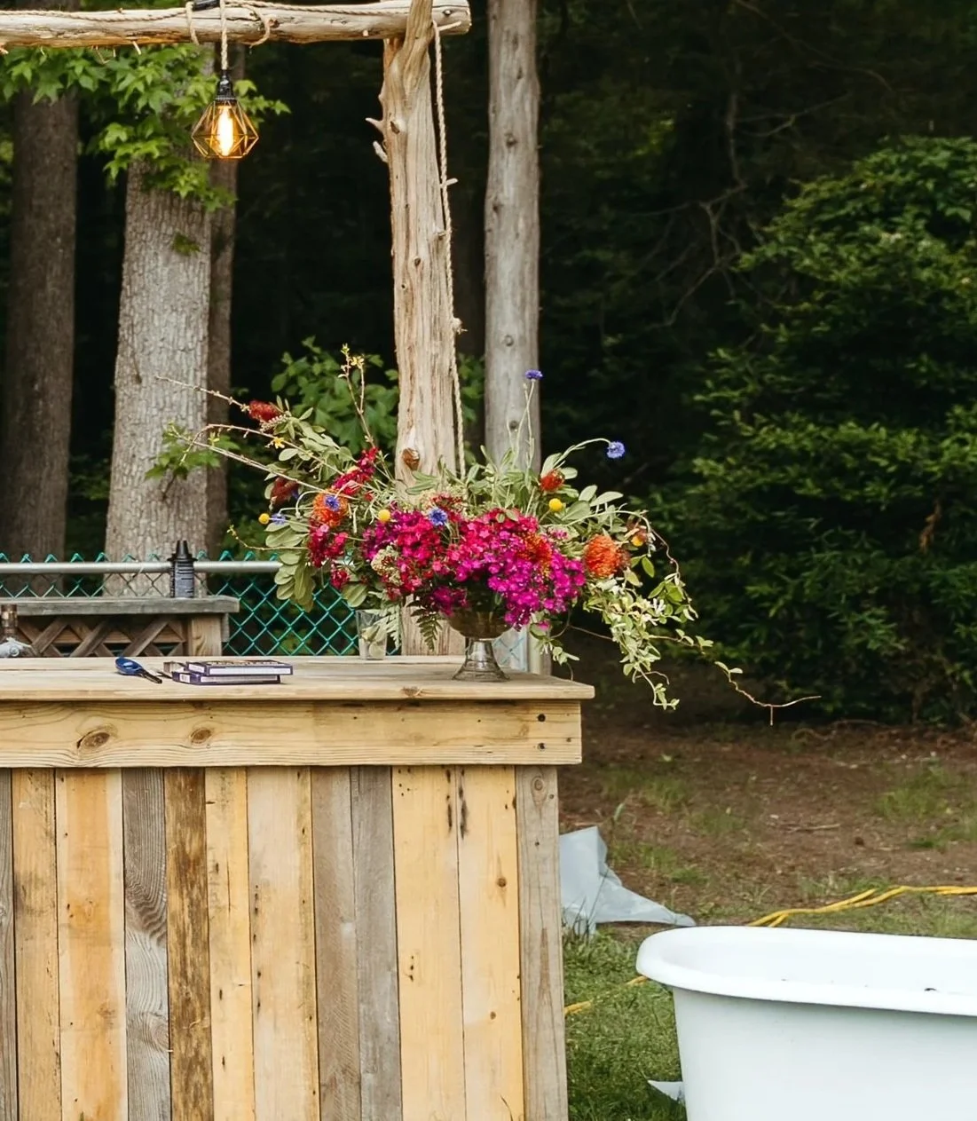 A wooden outdoor bar with a large flower arrangement on top, a hanging light fixture above, and a white clawfoot bathtub on the grass nearby, surrounded by trees.