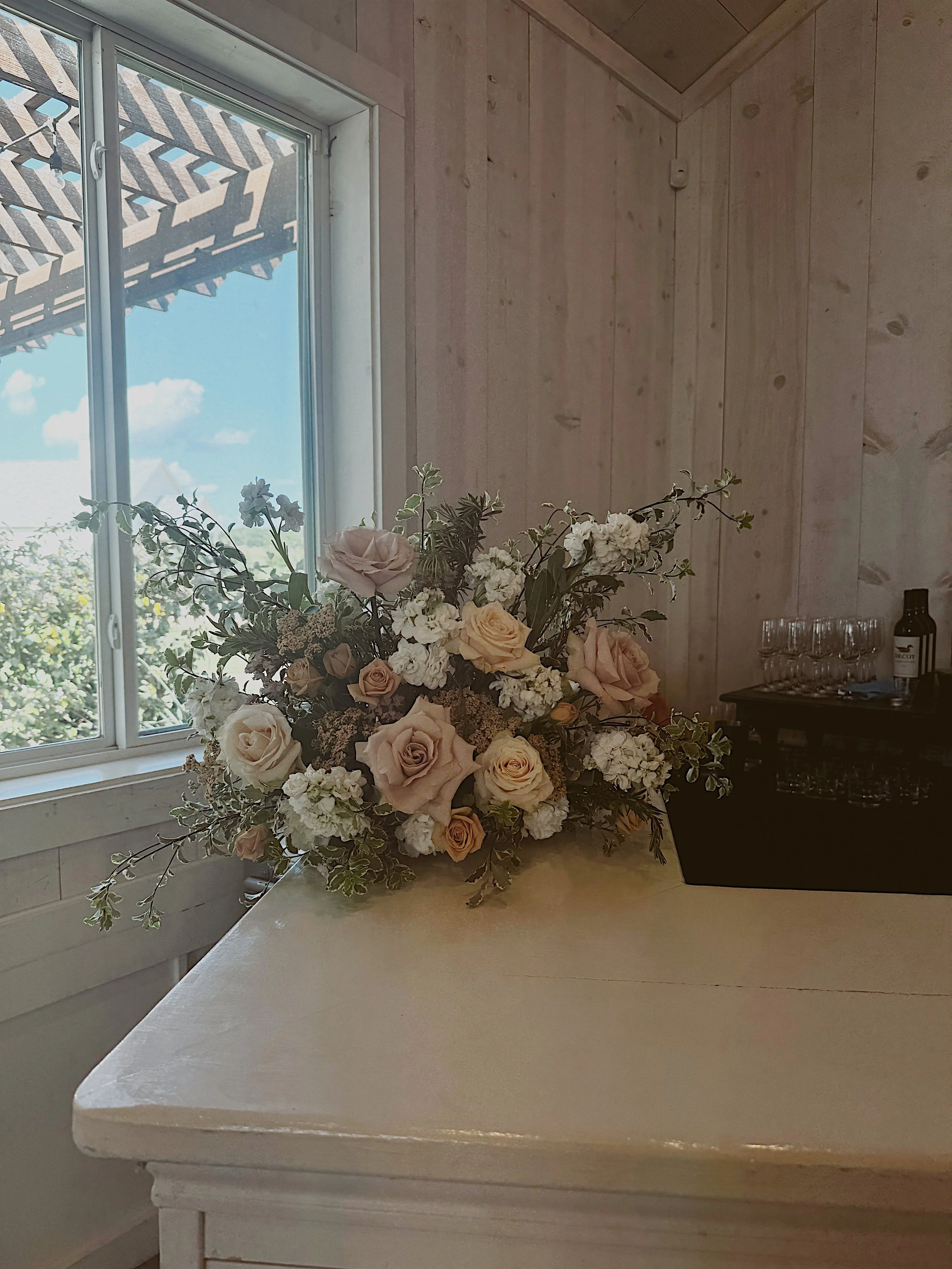 A large bouquet of light pink, white, and peach roses with greenery on a white table near a window showing a blue sky and trees outside.