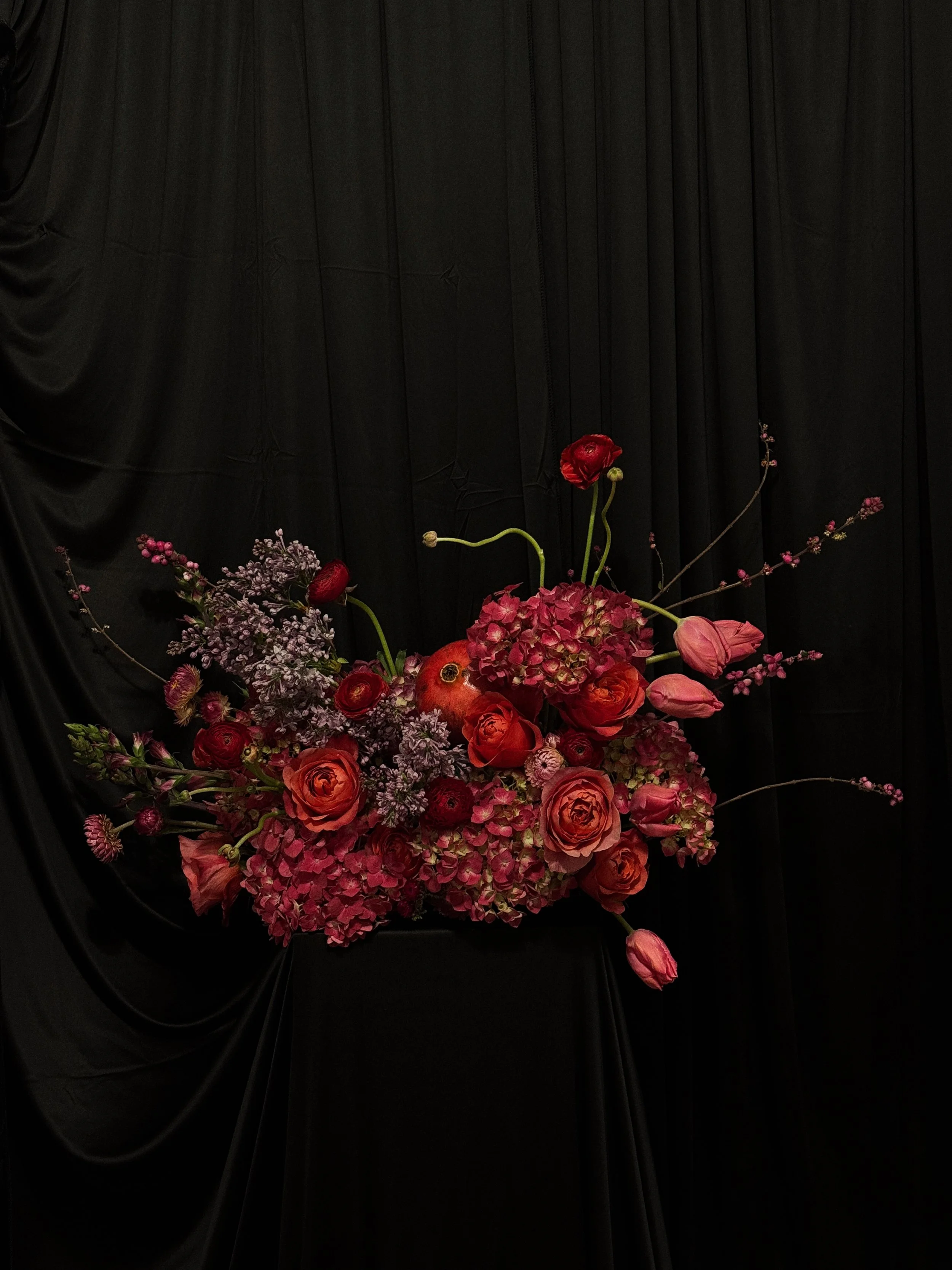 A floral arrangement with pink and red flowers, including roses and hydrangeas, set against a black fabric backdrop.