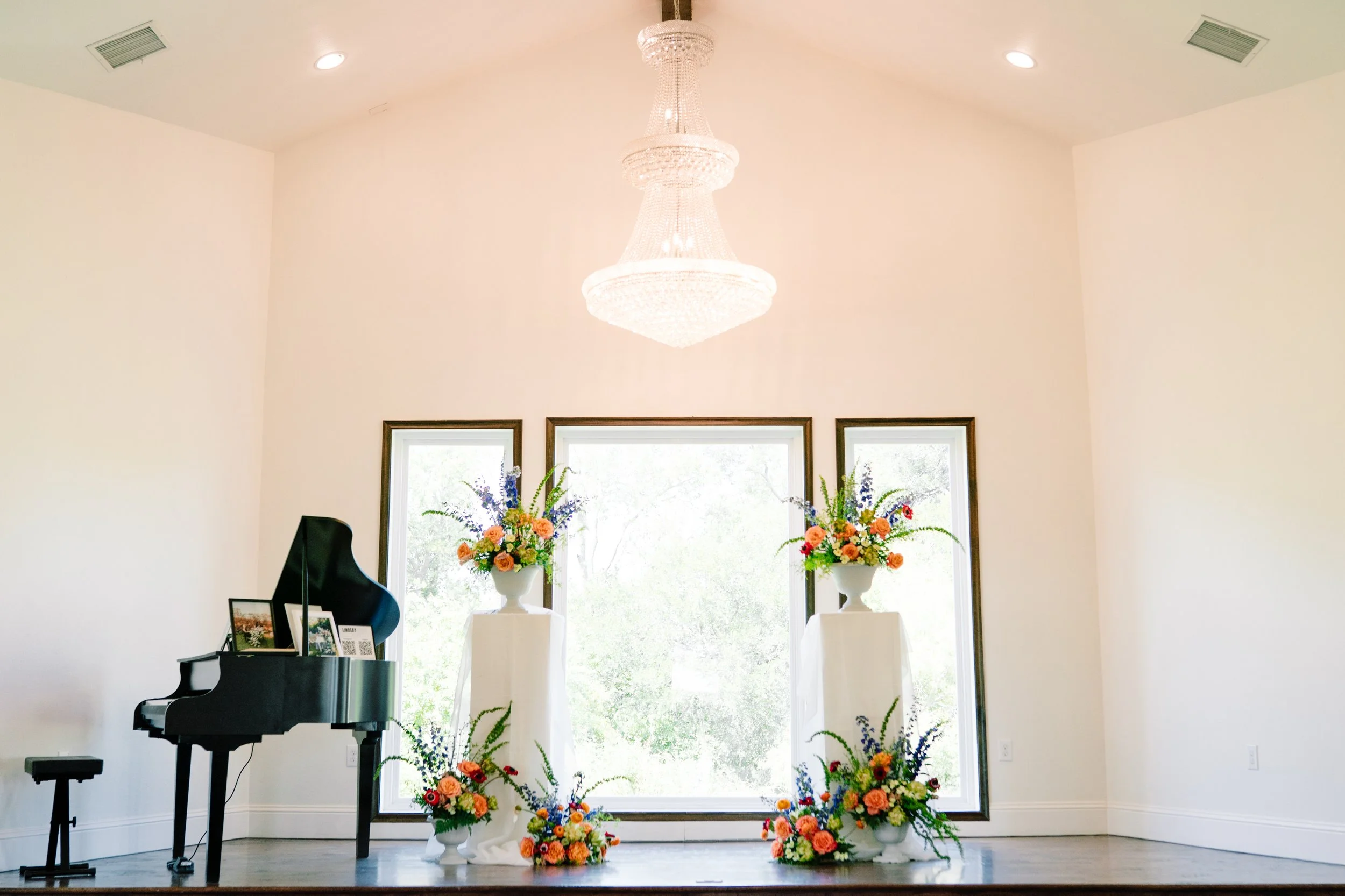 A room with a grand chandelier hanging from the ceiling, three windows in the background, two large floral arrangements on pedestals, a black grand piano with photos and a small framed picture on top, and three framed artworks on the wall.