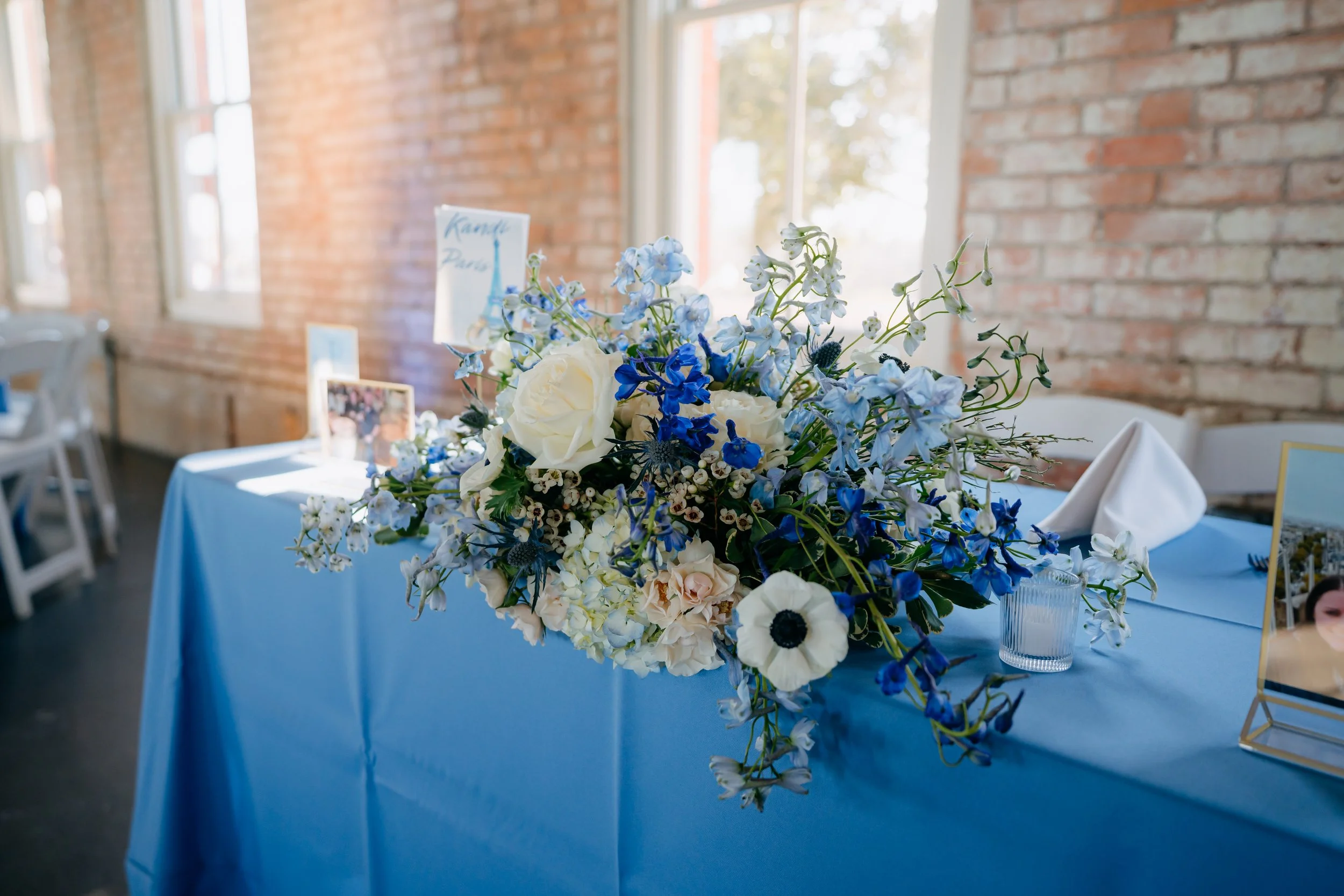A large bouquet of white and blue flowers, including roses, delphiniums, and anemones, arranged on a table with a blue tablecloth in a room with brick walls and large windows.