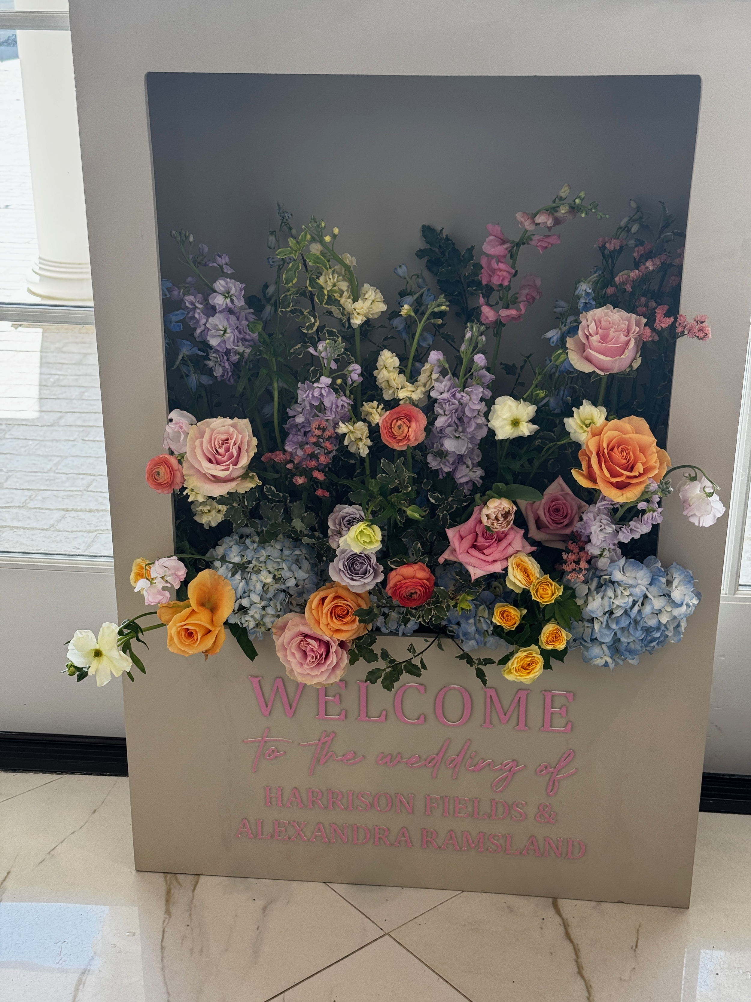 A floral arrangement with pink, yellow, purple, and white flowers on a gray welcome sign for a wedding at Harrison Fields and Alexandria Ramsland.