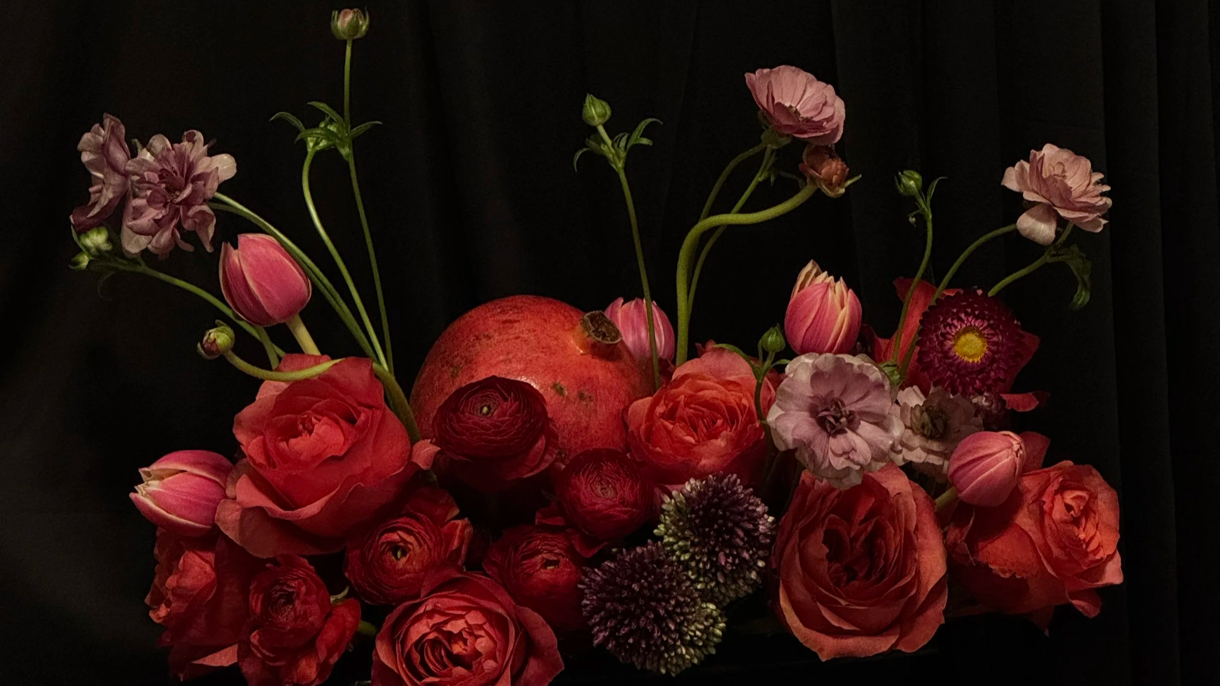 Arrangement of pink and red flowers and a pomegranate on a dark background.