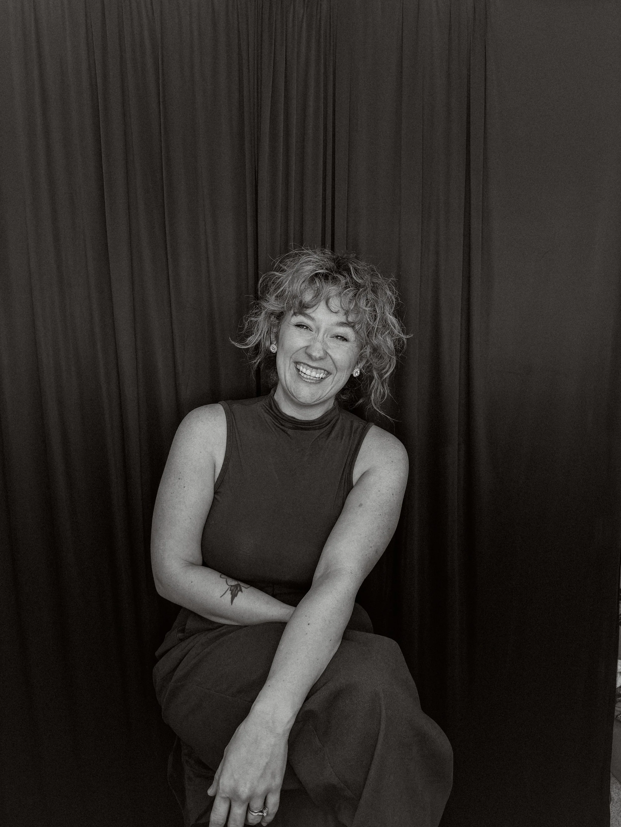 A smiling woman with curly hair sitting against a dark curtain backdrop.