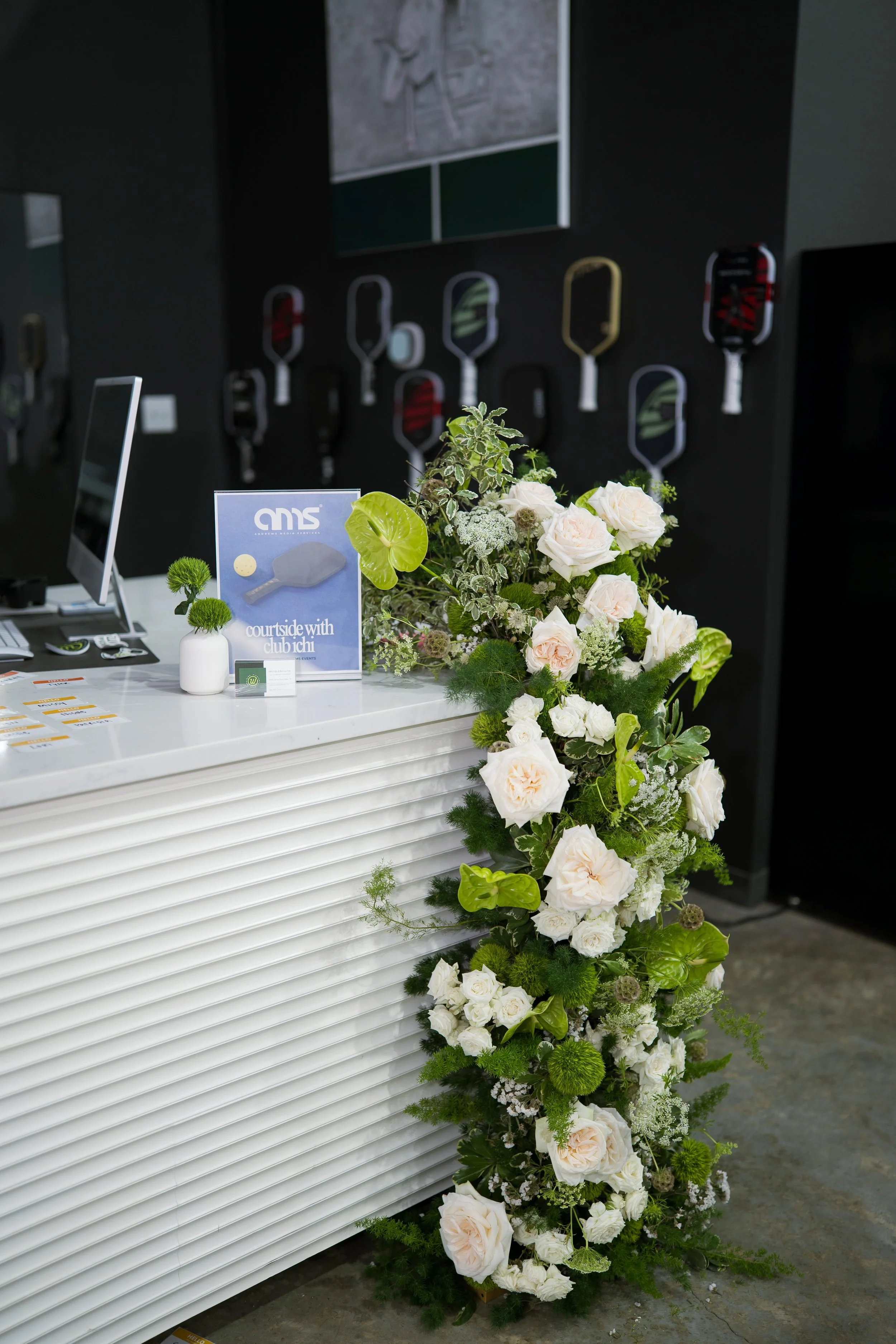 A floral arrangement with white roses, green leaves, and other white and green flowers on a white counter, with a black wall and paddle tennis rackets in the background.