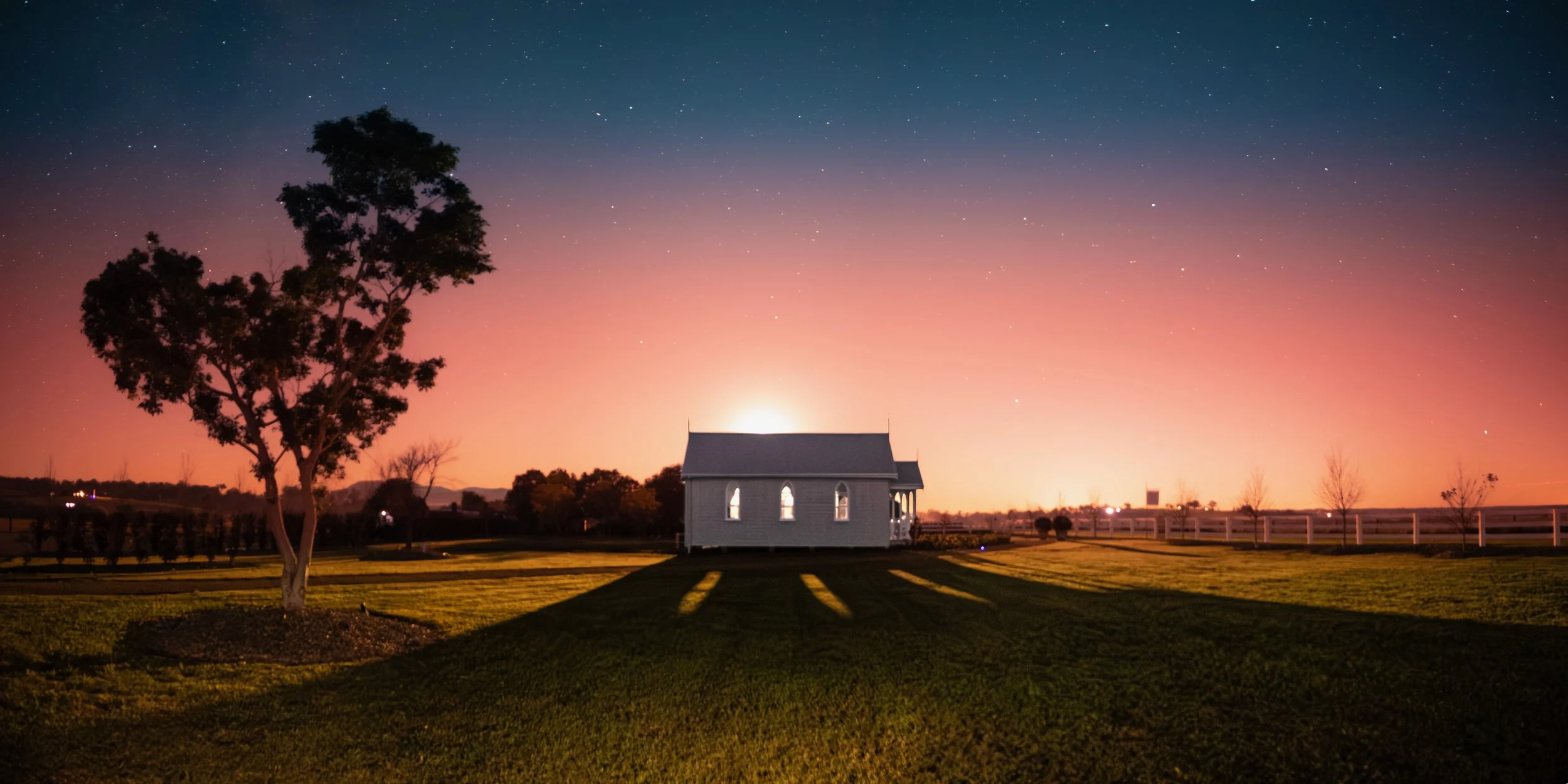A small white house with a porch and arched windows, located on a well-maintained lawn with landscaped flower beds. There is a large tree to the left of the house, casting shadows on the grass. The sky above is twilight with a gradient from pinkish-o