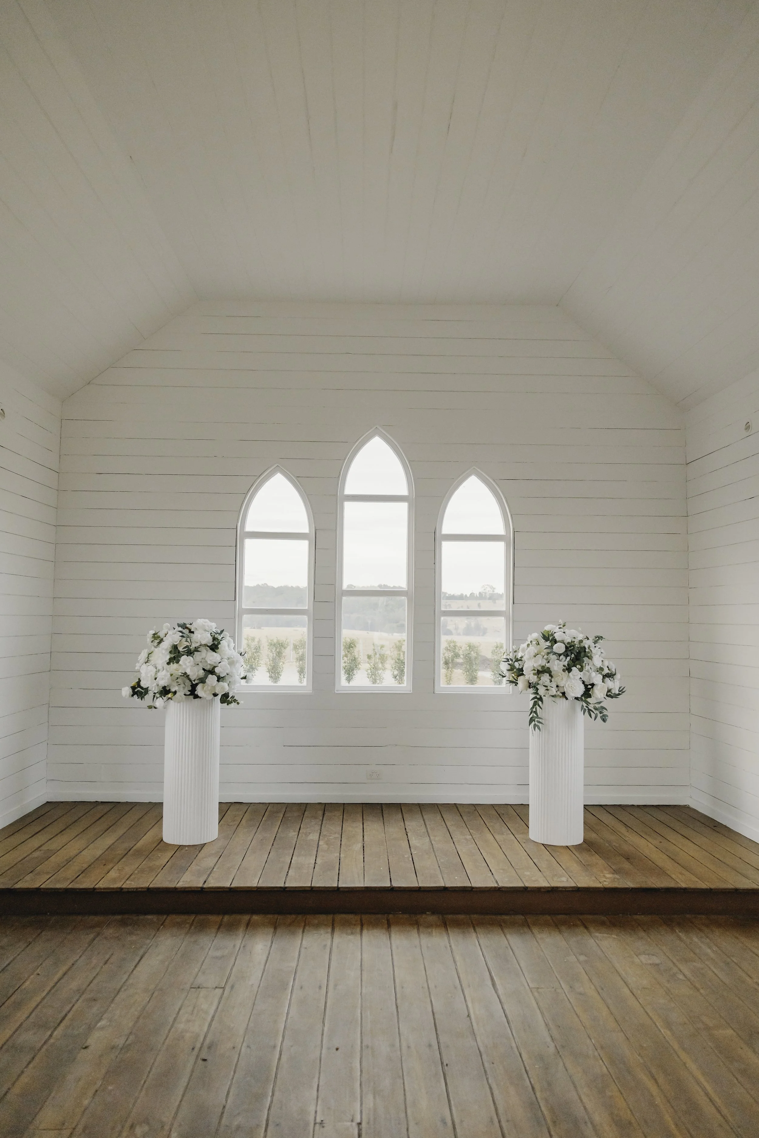 Interior of a white wooden chapel with three arched windows and two white pedestals holding floral arrangements.