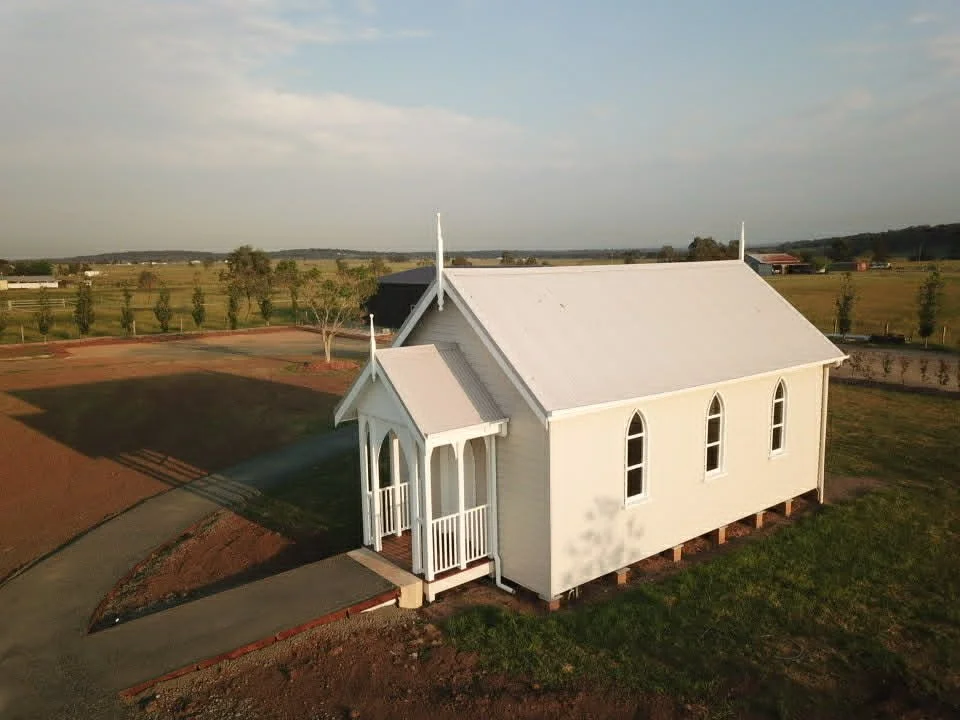 White church with arched windows and a small front porch, situated on a grassy area with a dirt and paved walkway, under a partly cloudy sky.