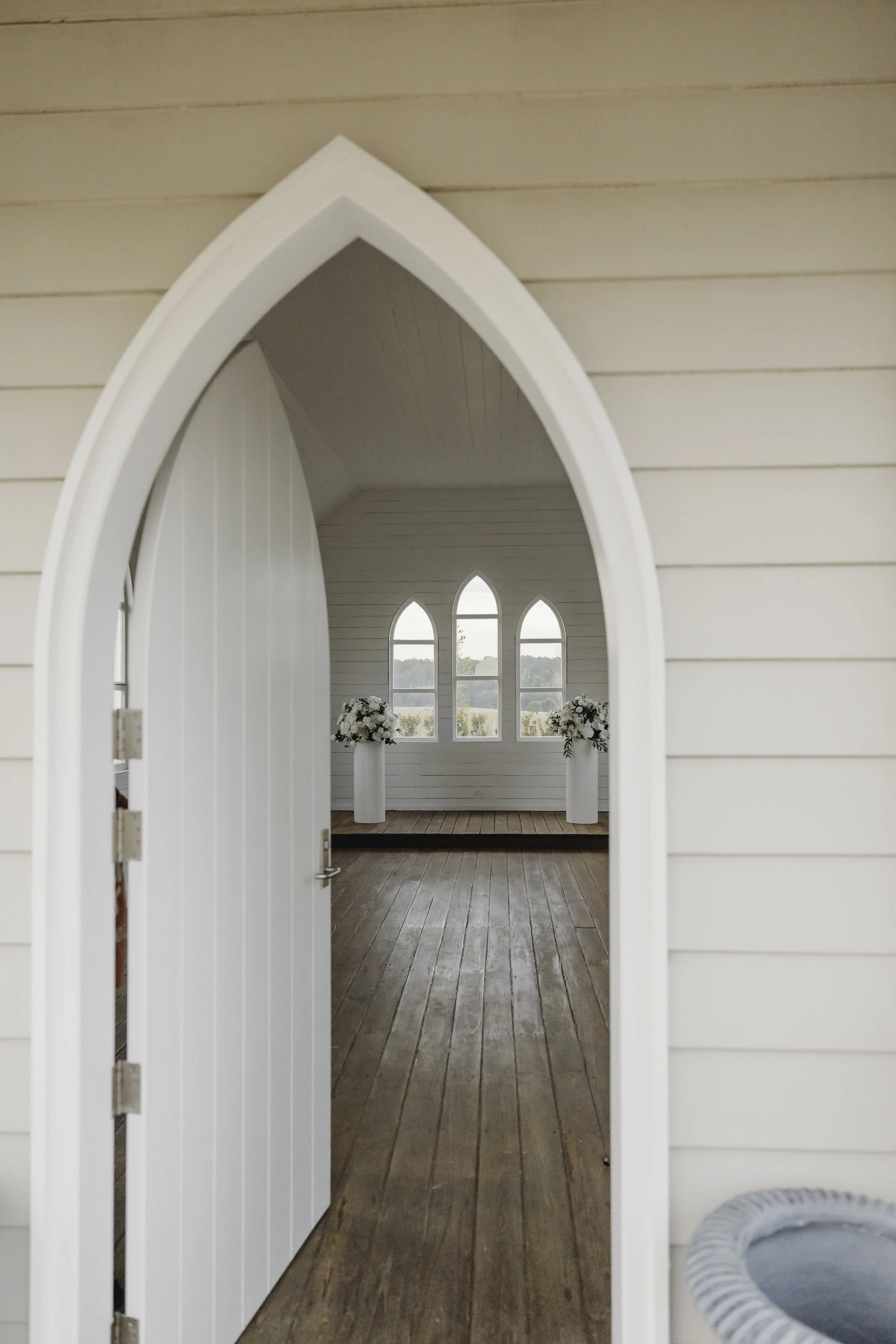 View through open white barn door into a room with arched windows, floral arrangements on pedestals, and wooden flooring.