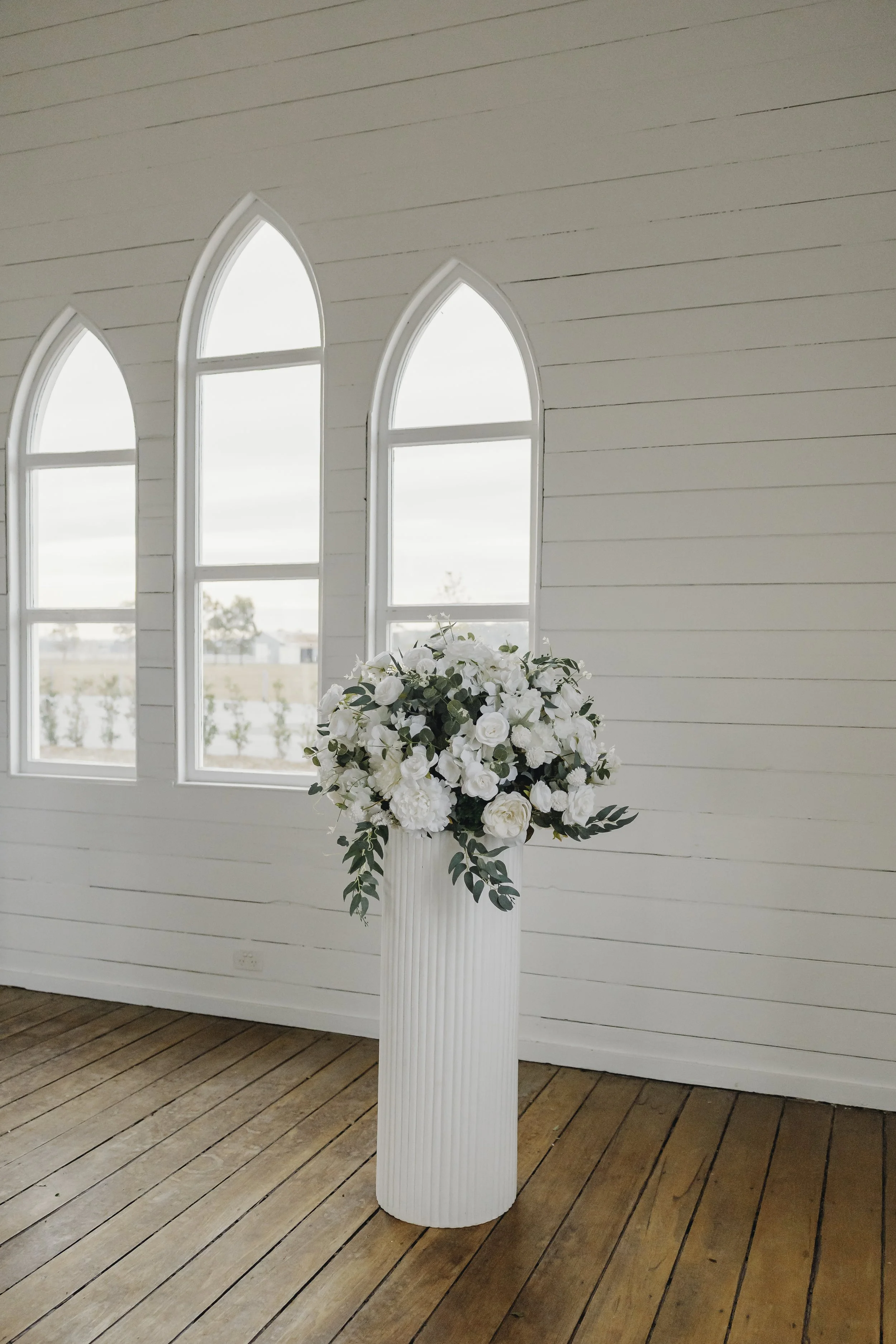 A white flower arrangement with flowers and green foliage in a tall, white, ribbed vase, placed in front of a white-washed wall with three arched windows.