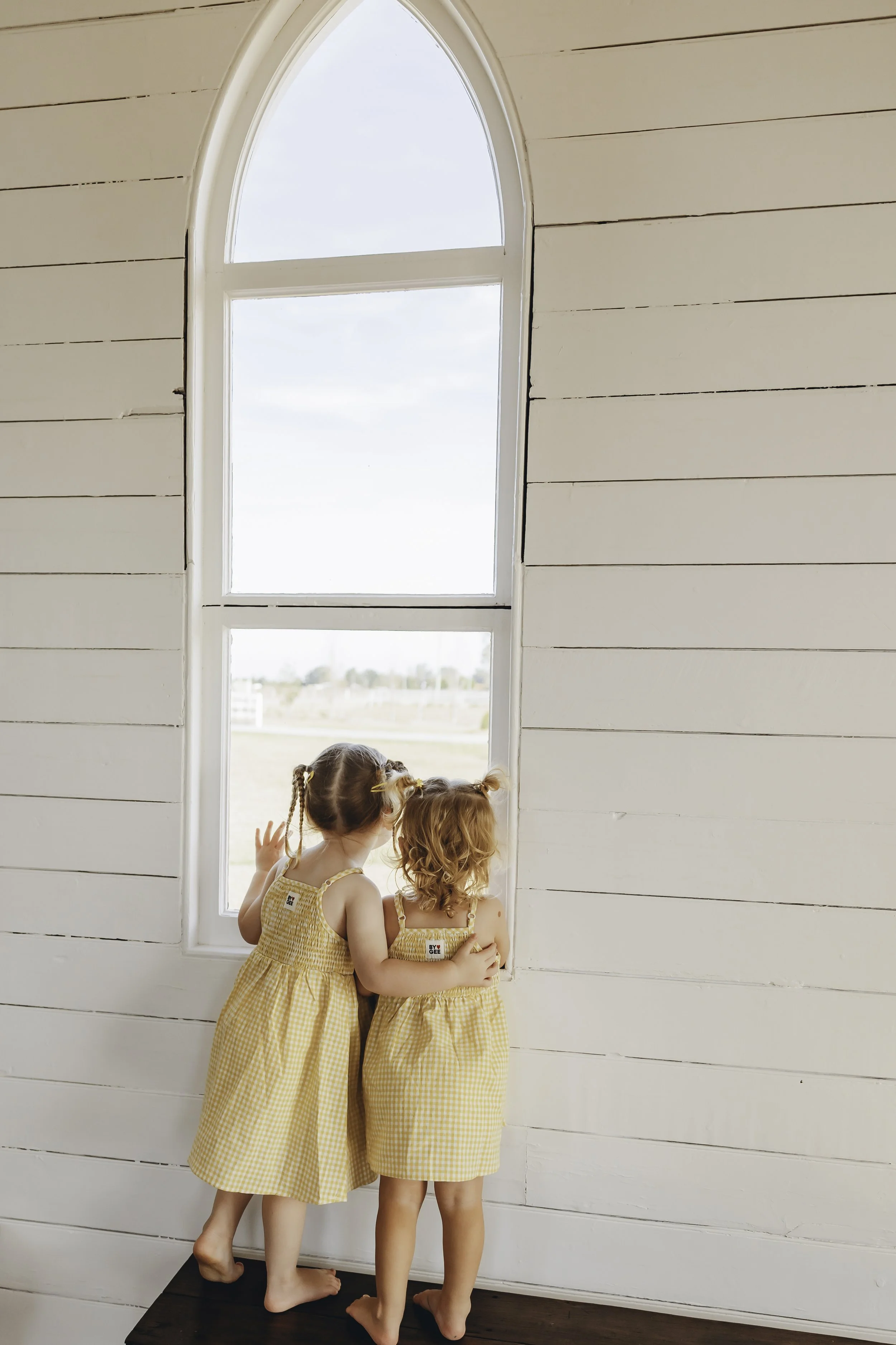 Two young girls with curly hair wearing matching yellow gingham dresses standing on a dark wooden step, looking out a large arched window in a room with white shiplap walls.
