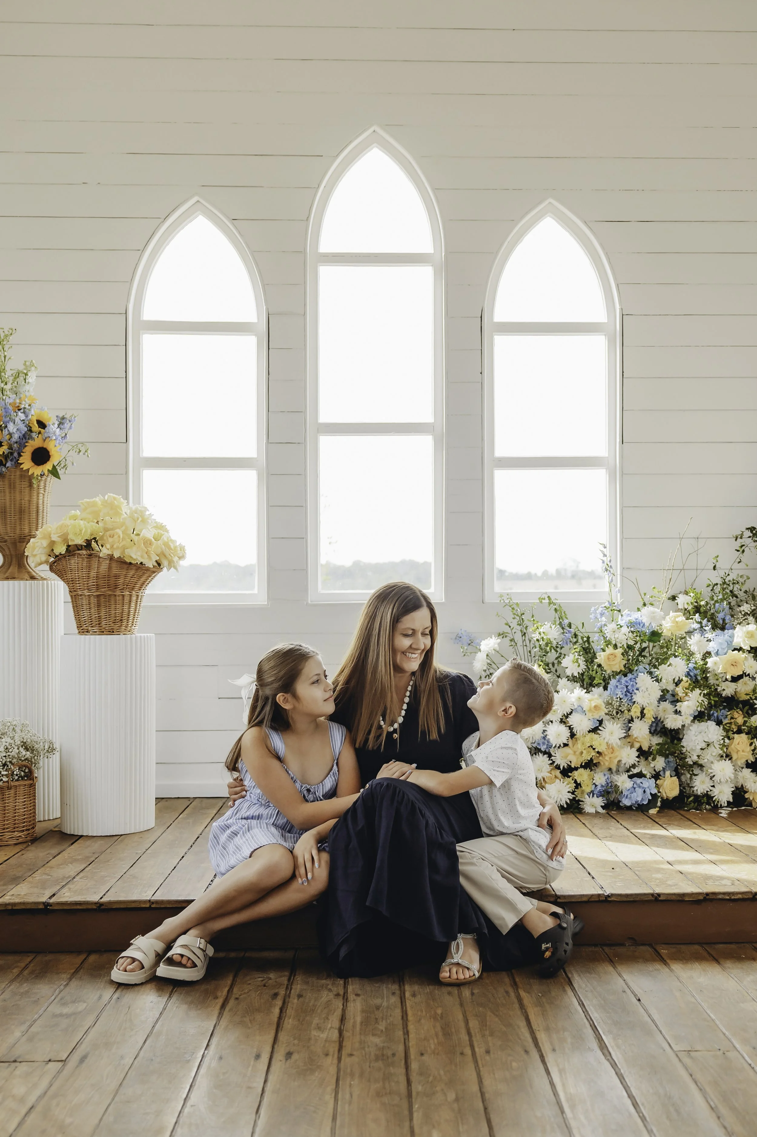 A woman and three children sitting on a wooden floor in front of a large, bright window with arched panes, surrounded by floral arrangements, with sunlight streaming in.