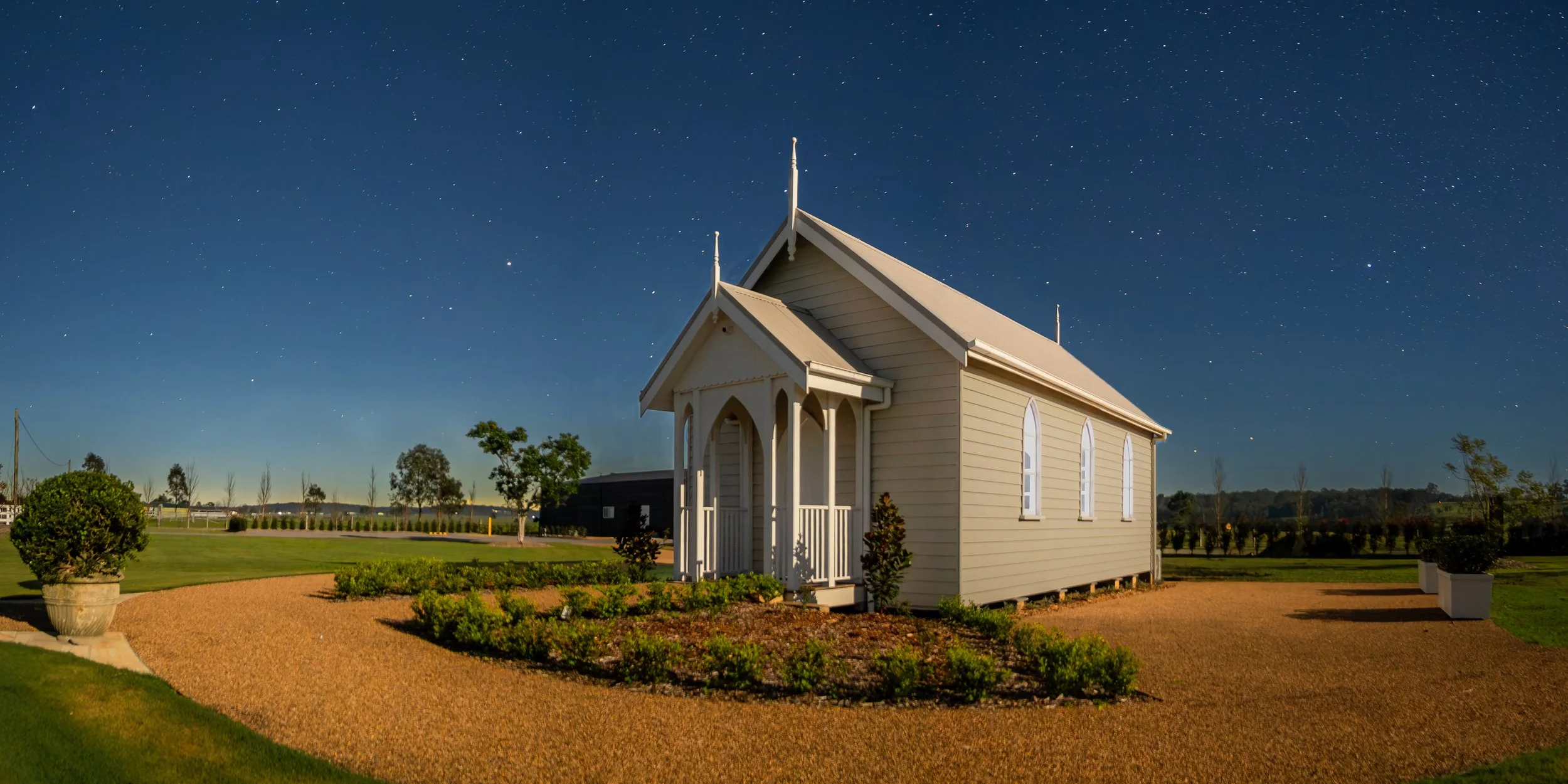 Small white church building under a starry night sky, surrounded by trees, bushes, and a grassy area.